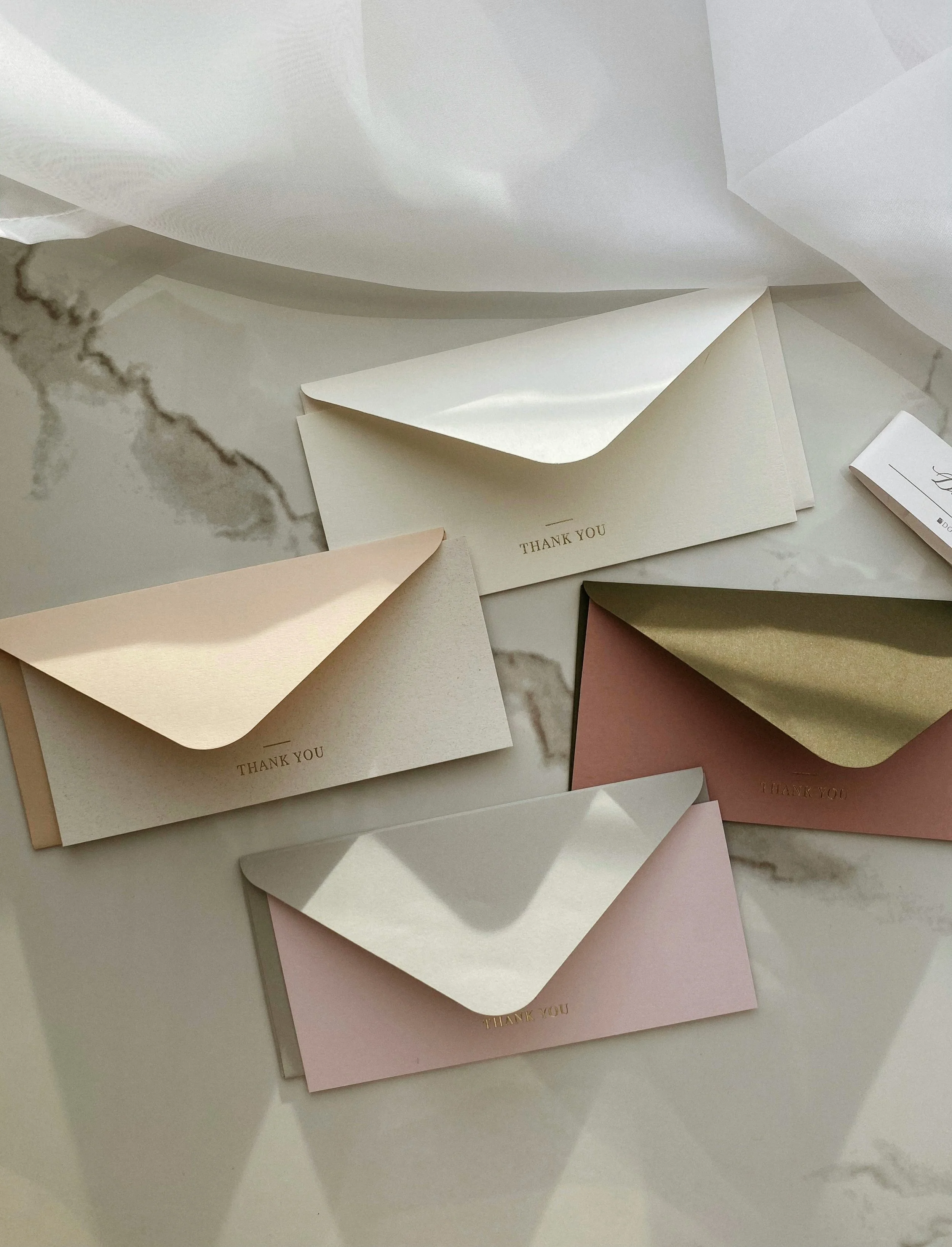 Four pastel-colored envelopes with 'Thank You' printed on them, placed on a marble surface with soft lighting.