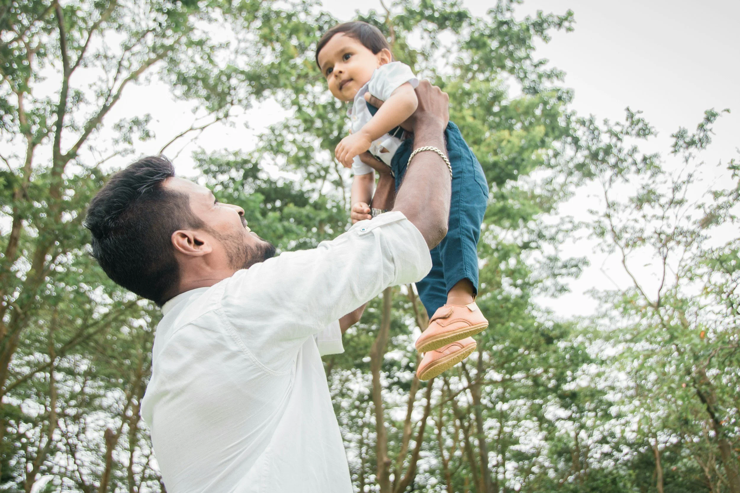 A man lifting a young boy in the air outdoors among trees.