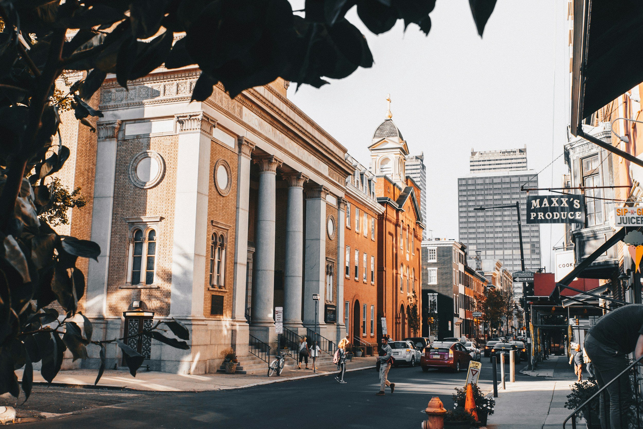A city street scene with a historic building featuring large columns, a church steeple, and modern high-rise buildings in the background. People are walking, and cars are parked along the street.
