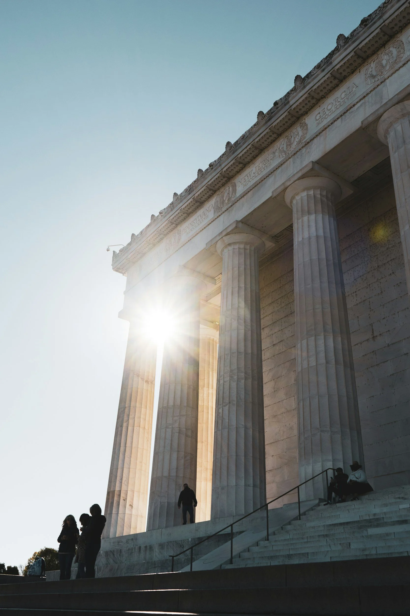 Looking up at the front of the Lincoln Memorial, showing columns with the sun shining between them, and people sitting and standing on the steps.