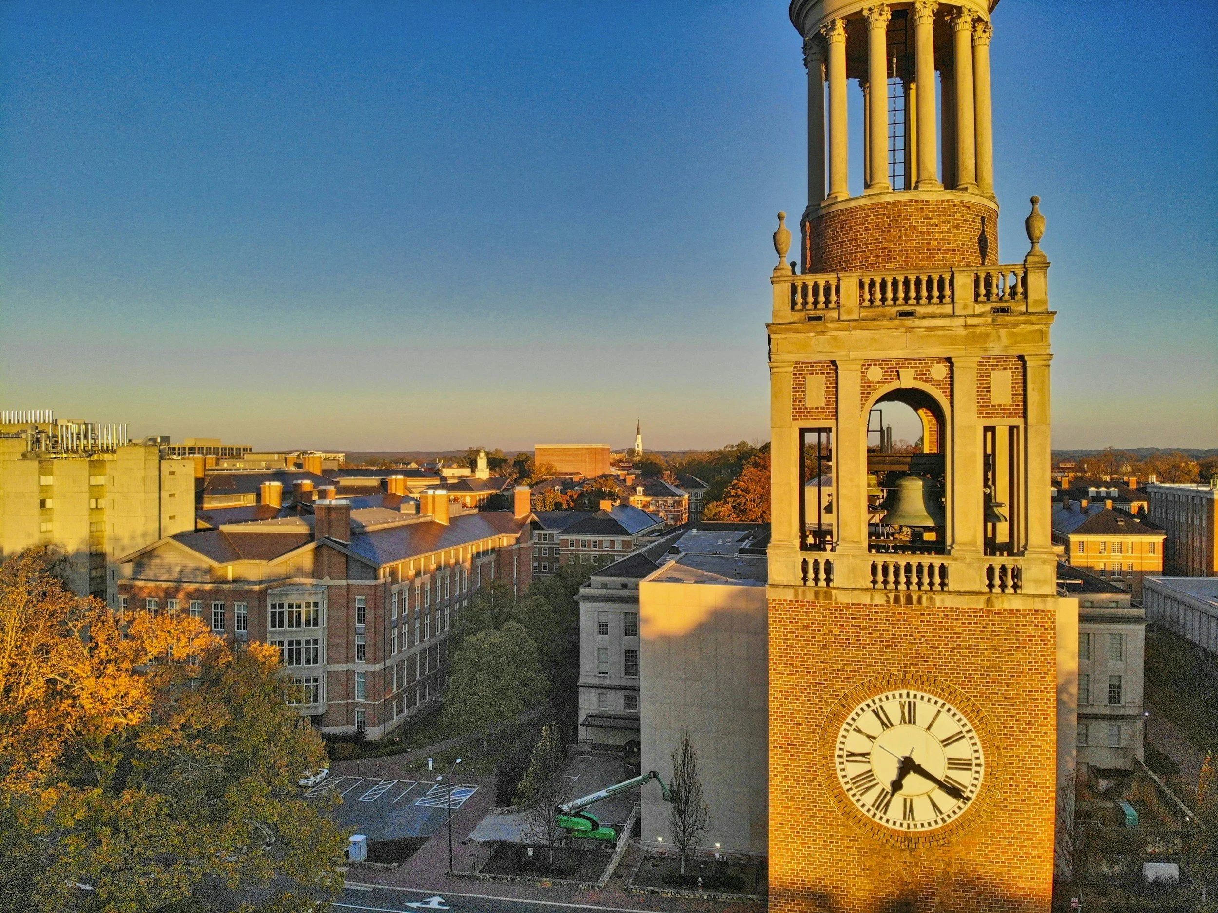 A historic clock tower with a large clock face showing the time as 4:20, located in an urban area during sunset, with surrounding buildings, trees, and a clear blue sky.