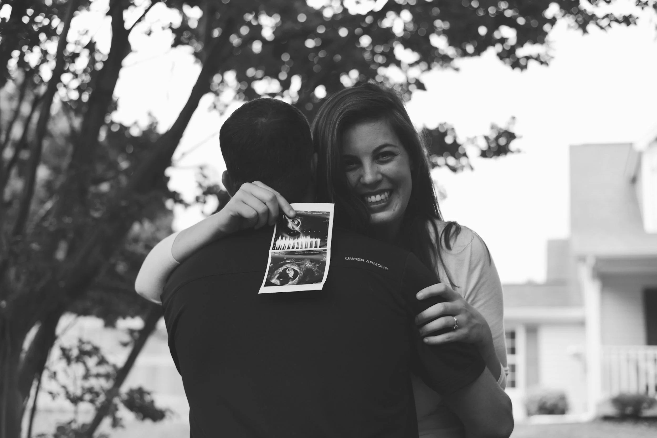 A woman smiling and hugging a man with an ultrasound photo attached to his shirt outside near trees and houses.