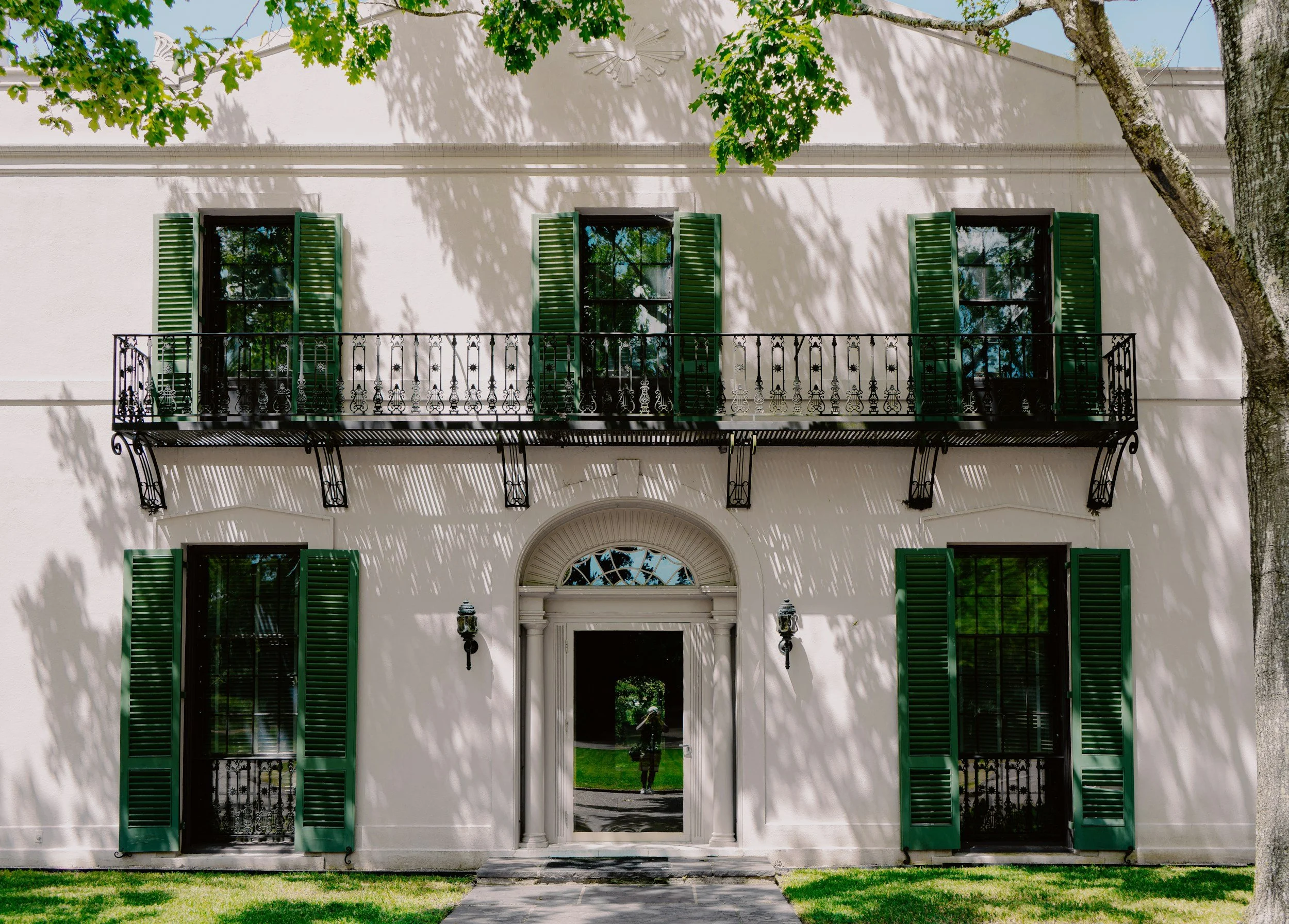 A white two-story house with a central entrance door, six windows with green shutters, and a black wrought iron balcony on the second floor. The front yard has green grass and a large tree casting shadows on the house.