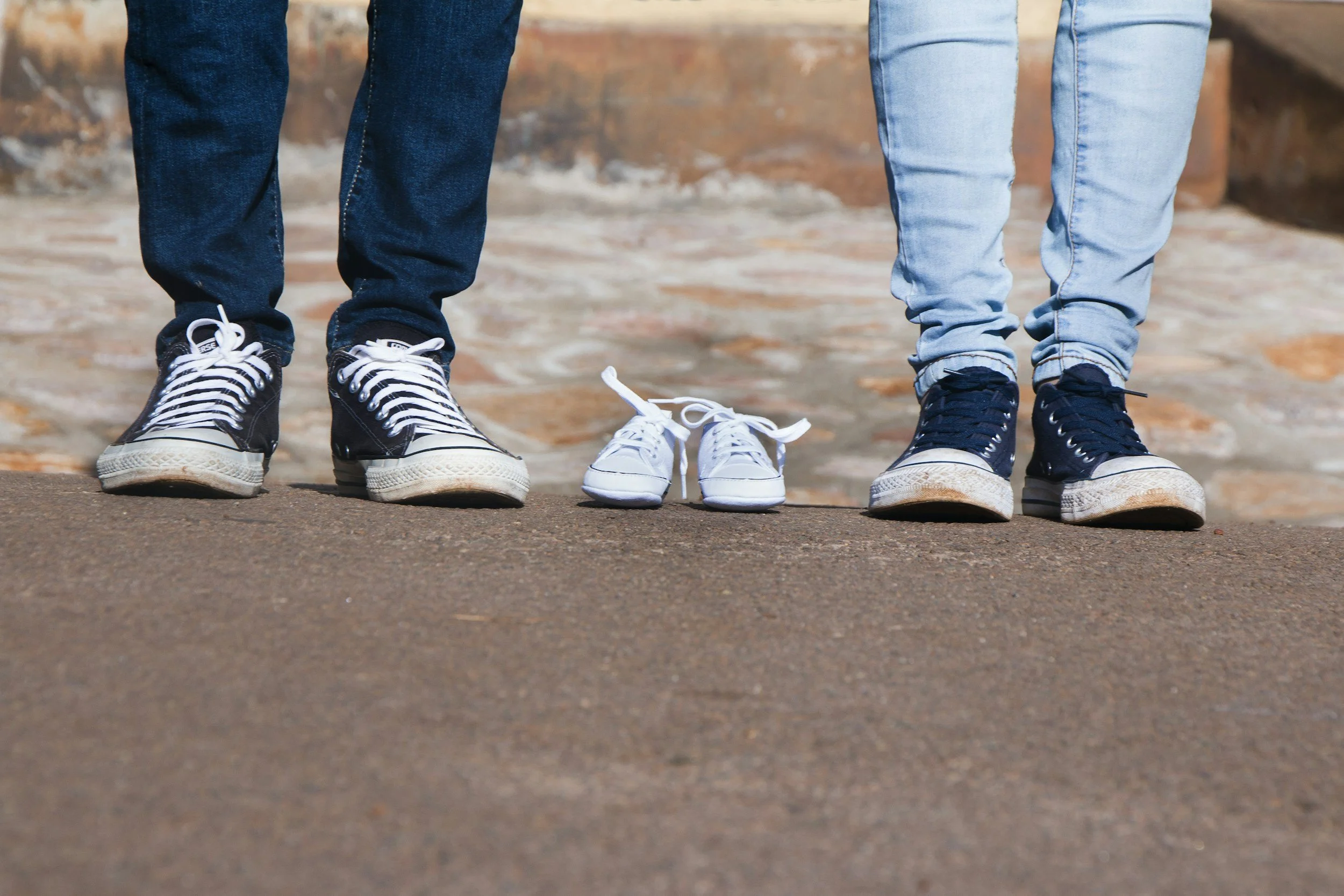 A pair of teenagers and two pairs of small white baby shoes on the ground, with only the lower legs and shoes visible in the image.