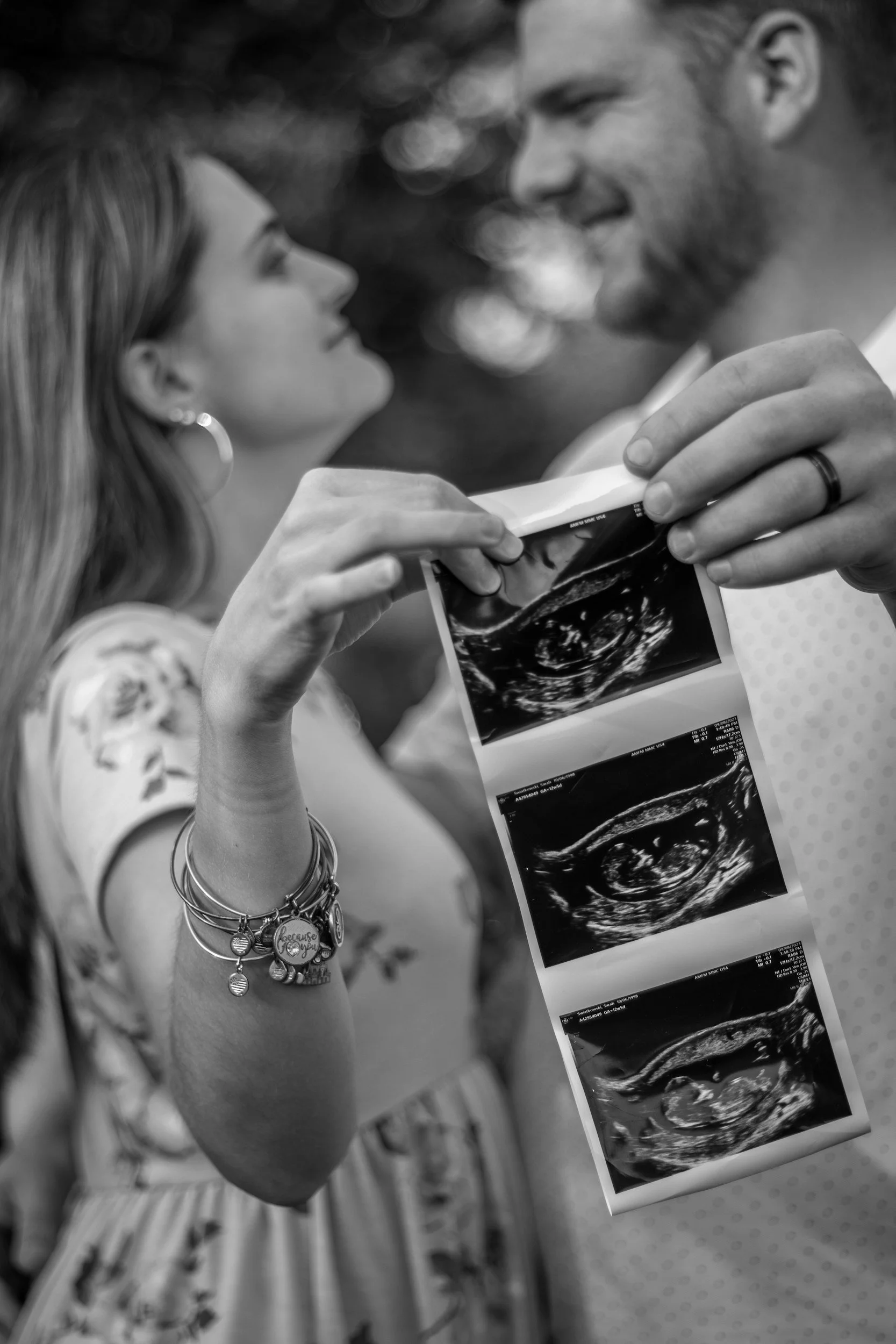 Couple holding ultrasound pictures, the woman with jewelry on her wrist, smiling and looking at the man, in black and white photo.