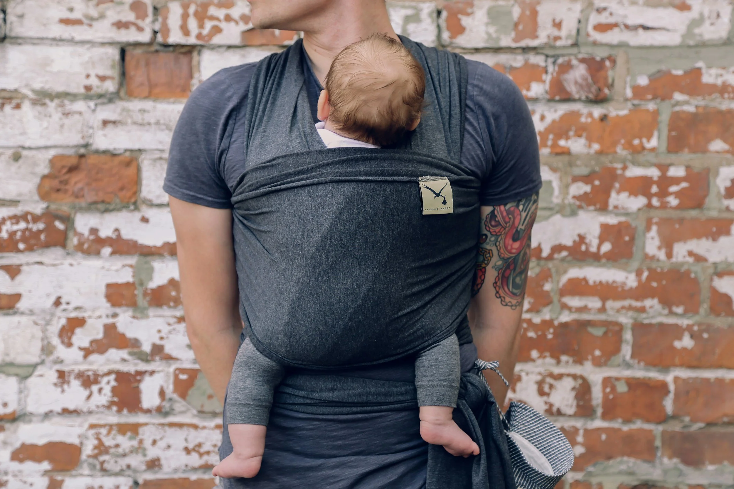 A person wearing a black t-shirt is carrying a baby in a black baby carrier against a brick wall background.