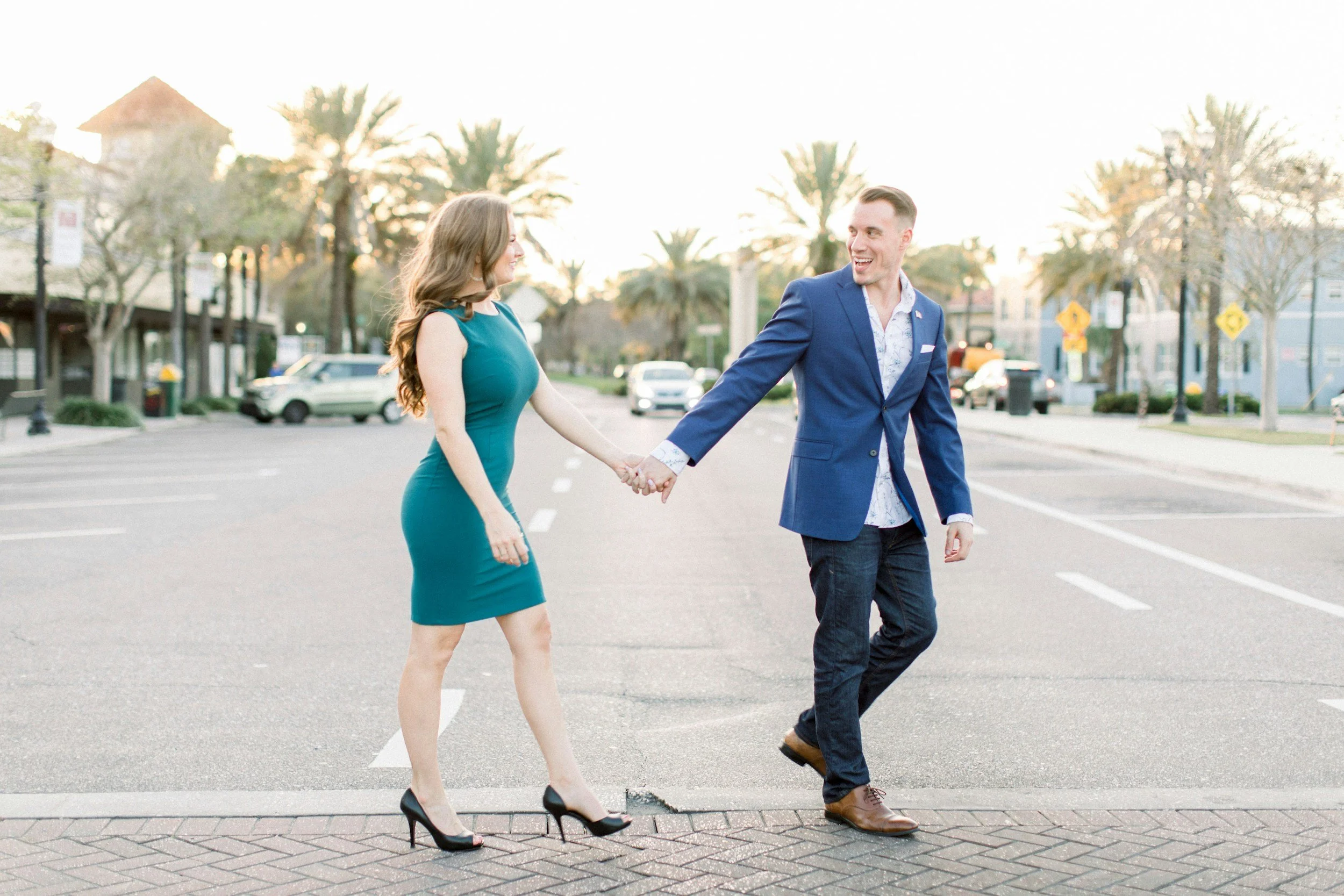 A man and woman holding hands and walking across a city street at sunset, smiling at each other.