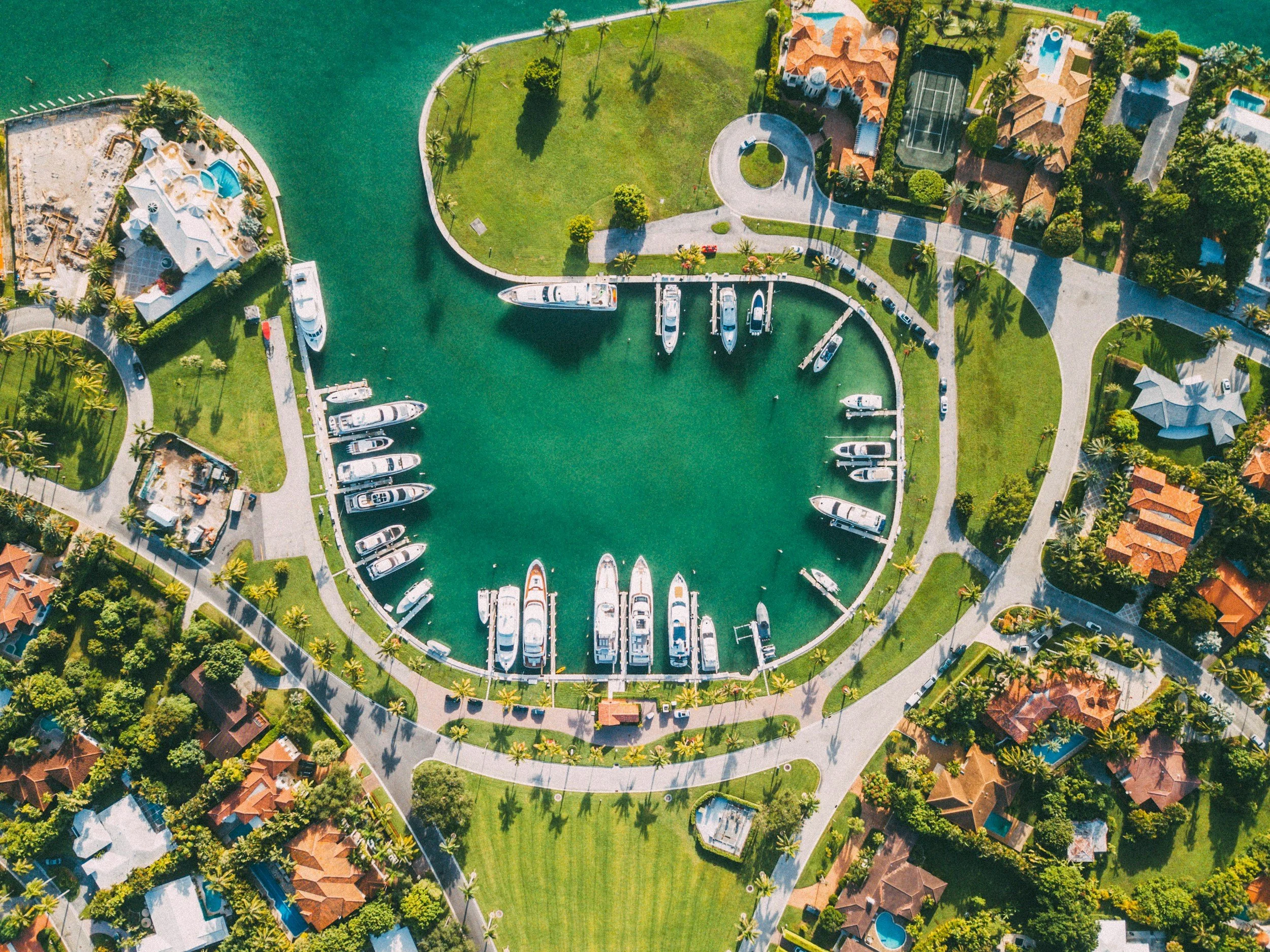 Aerial view of a marina with boats docked in water surrounded by green lawns, residential houses, and paved roads.