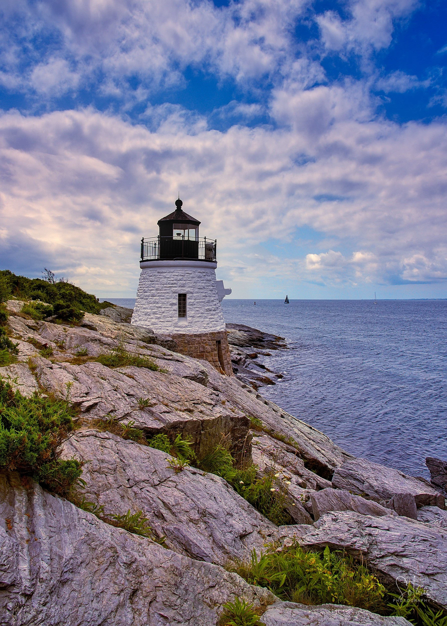 A white lighthouse on rocky coastline with a cloudy sky and sailboats in the distance.