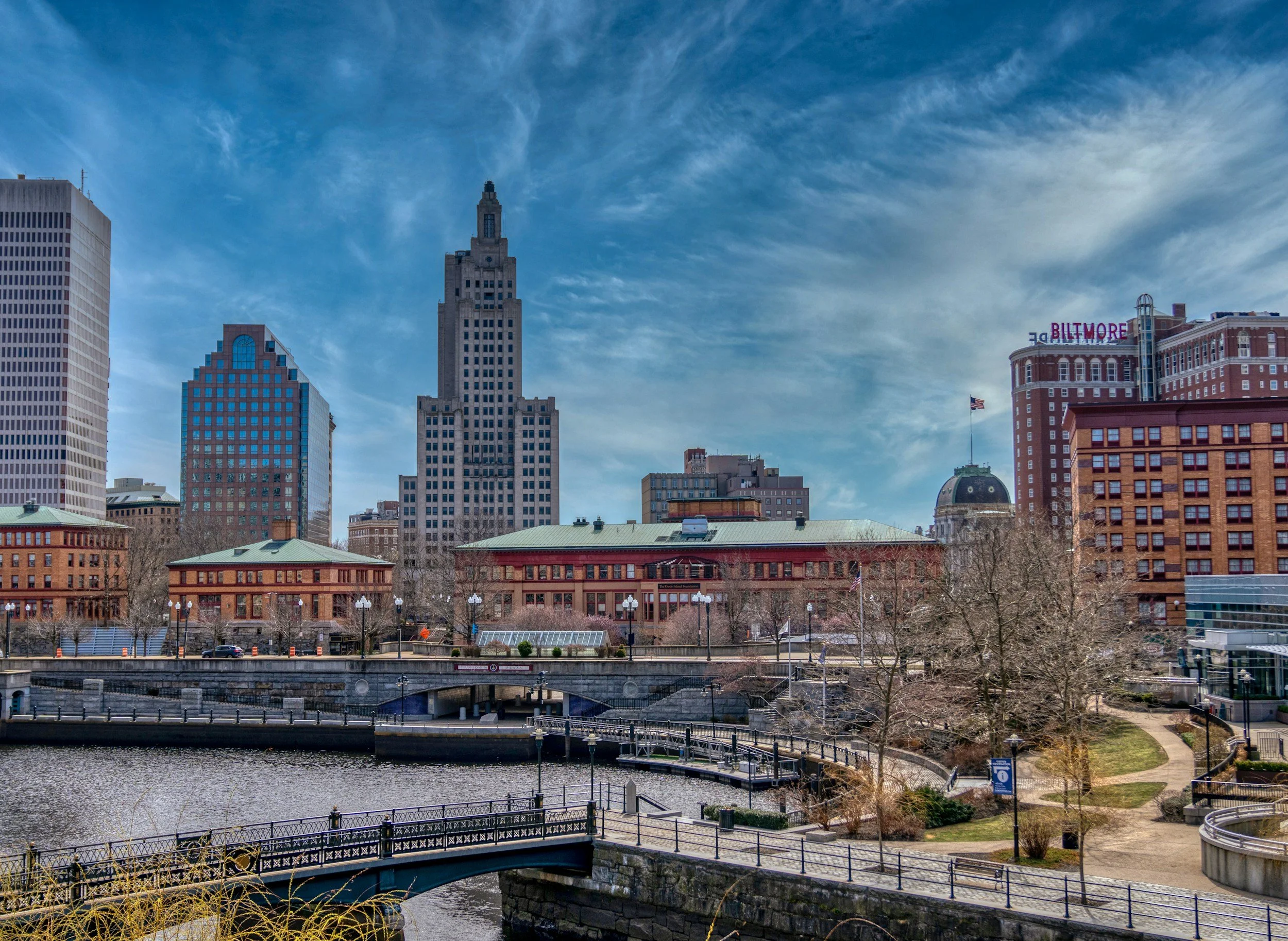 City skyline with tall buildings, a river, a park, and a bridge in the foreground under a partly cloudy sky.