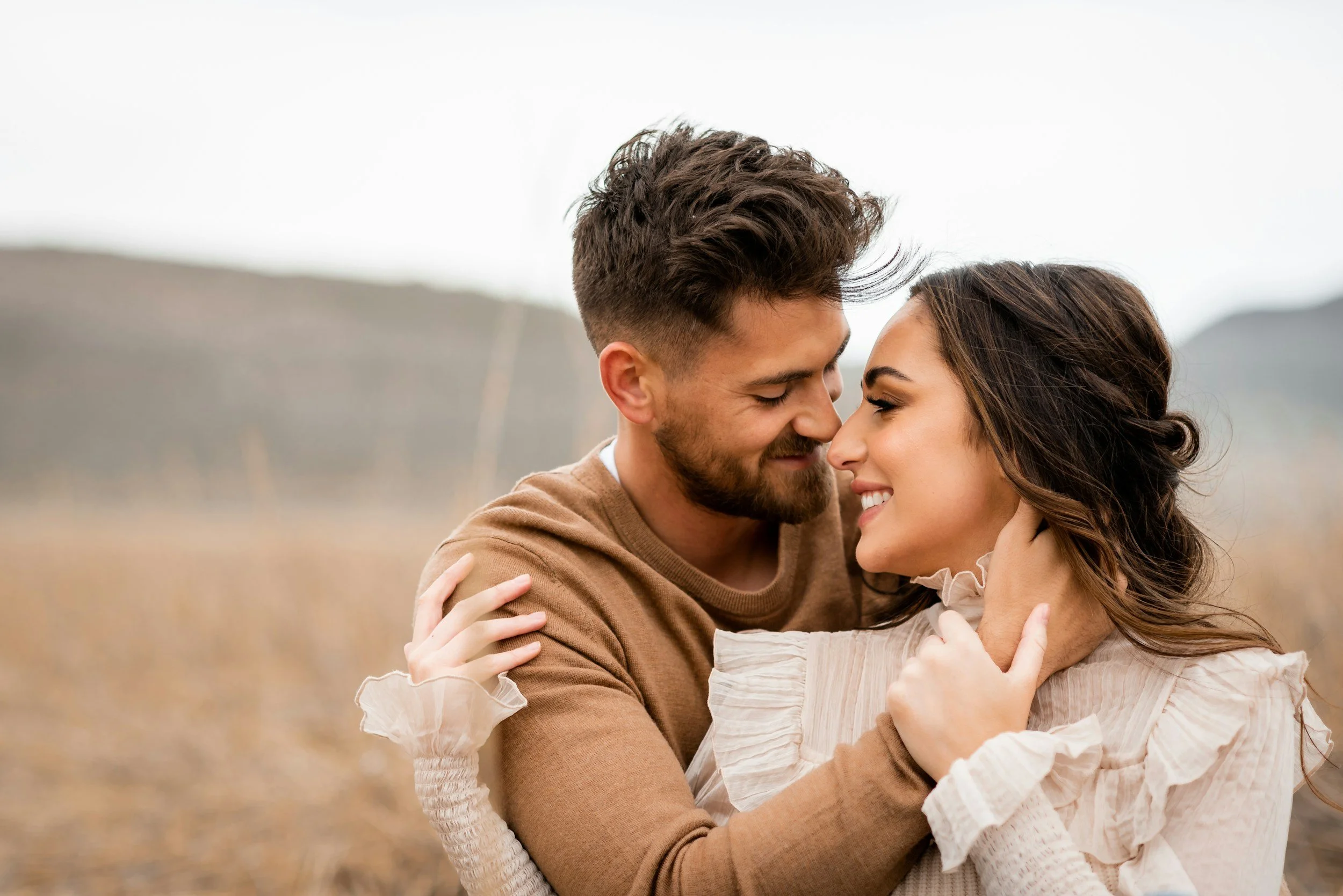 A couple embraces outdoors in a field, smiling and touching foreheads, with hills in the background.