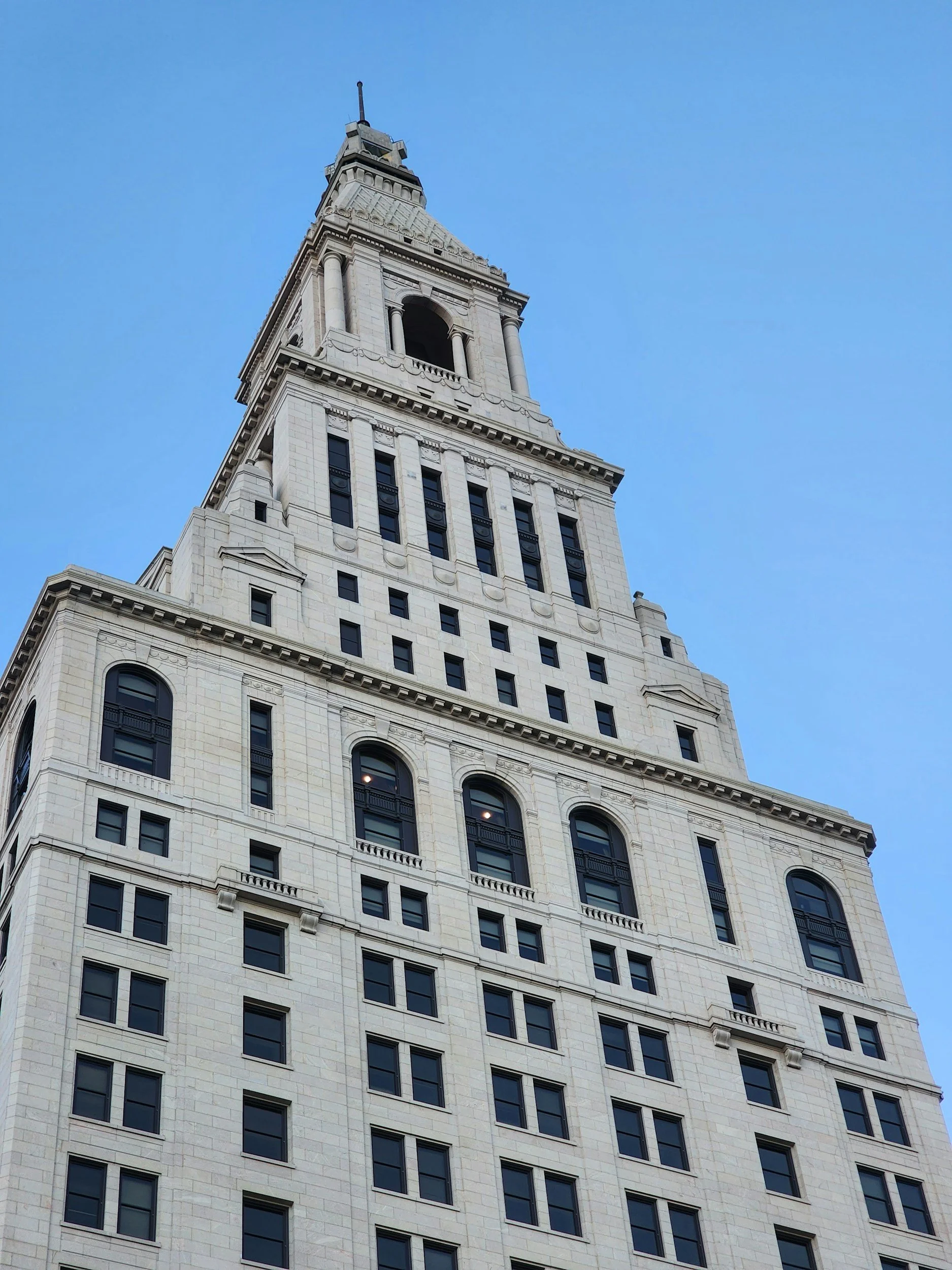 A tall historic skyscraper with a white stone facade, multiple arched windows, and a clock tower at the top, set against a clear blue sky.