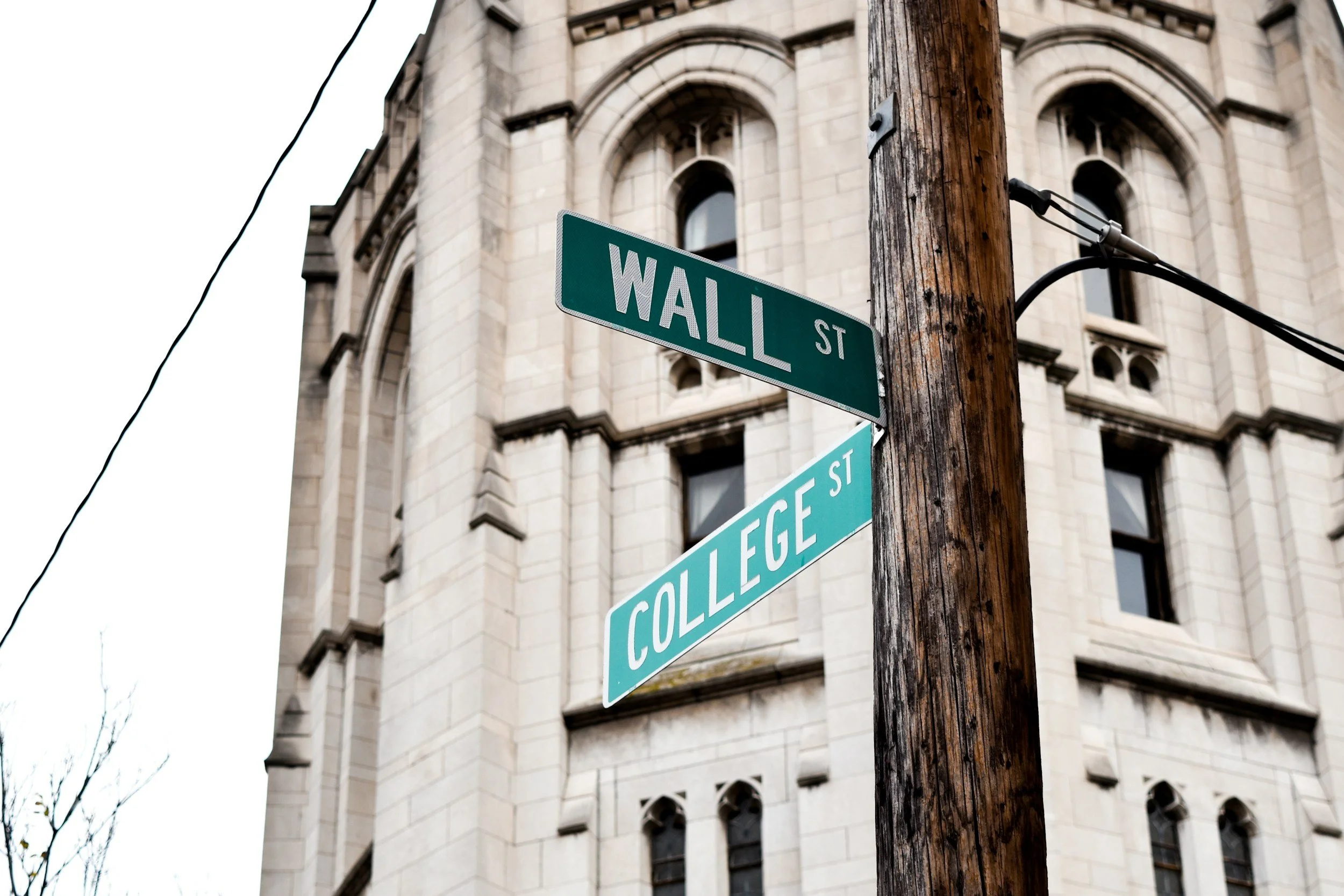 Street signs for Wall Street and College Street at an intersection in front of a historic building with arched windows.