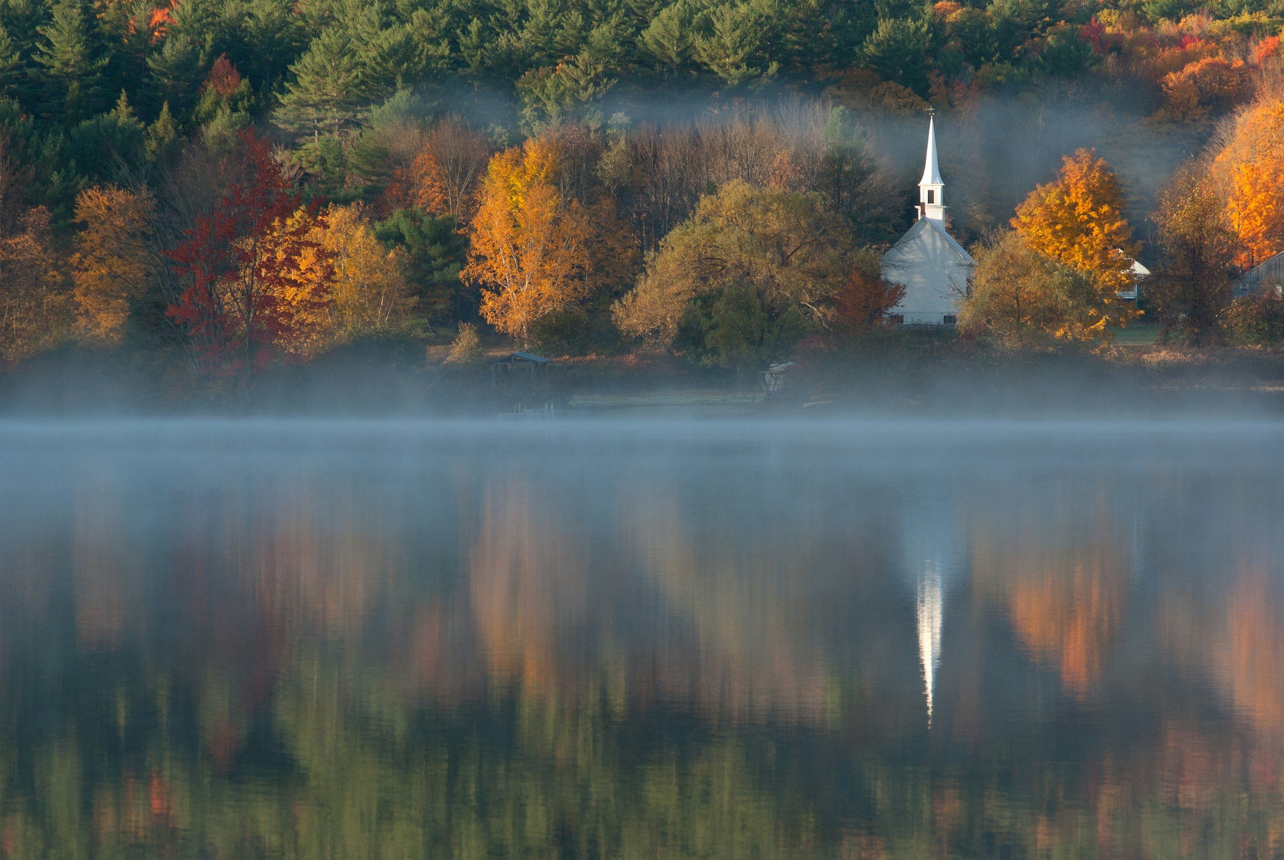 A peaceful lakeside scene with autumn foliage, fog rolling over the water, and a white church with a tall steeple among colorful trees on the hillside.