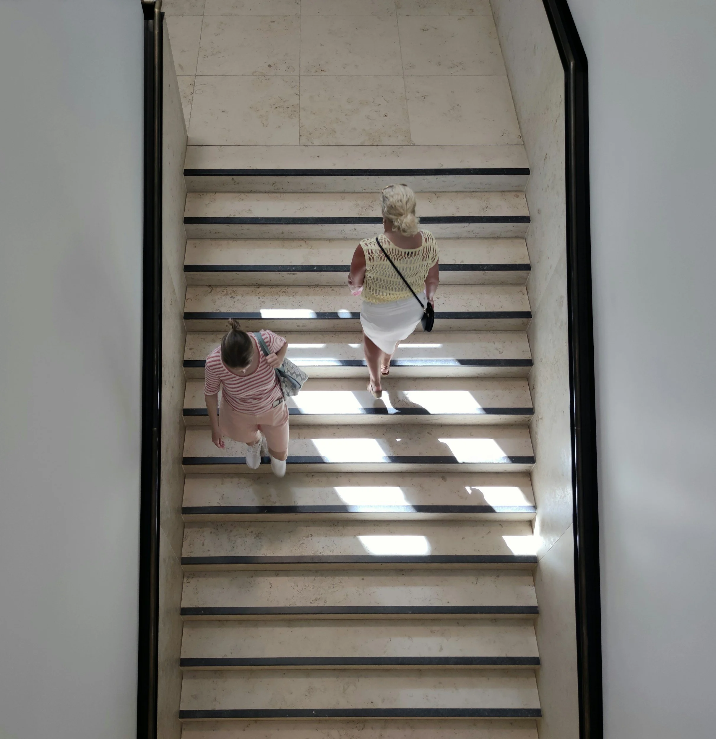 View from above of a woman and a girl walking up marble stairs with black edge trim, sunlight casting shadows on the stairs.