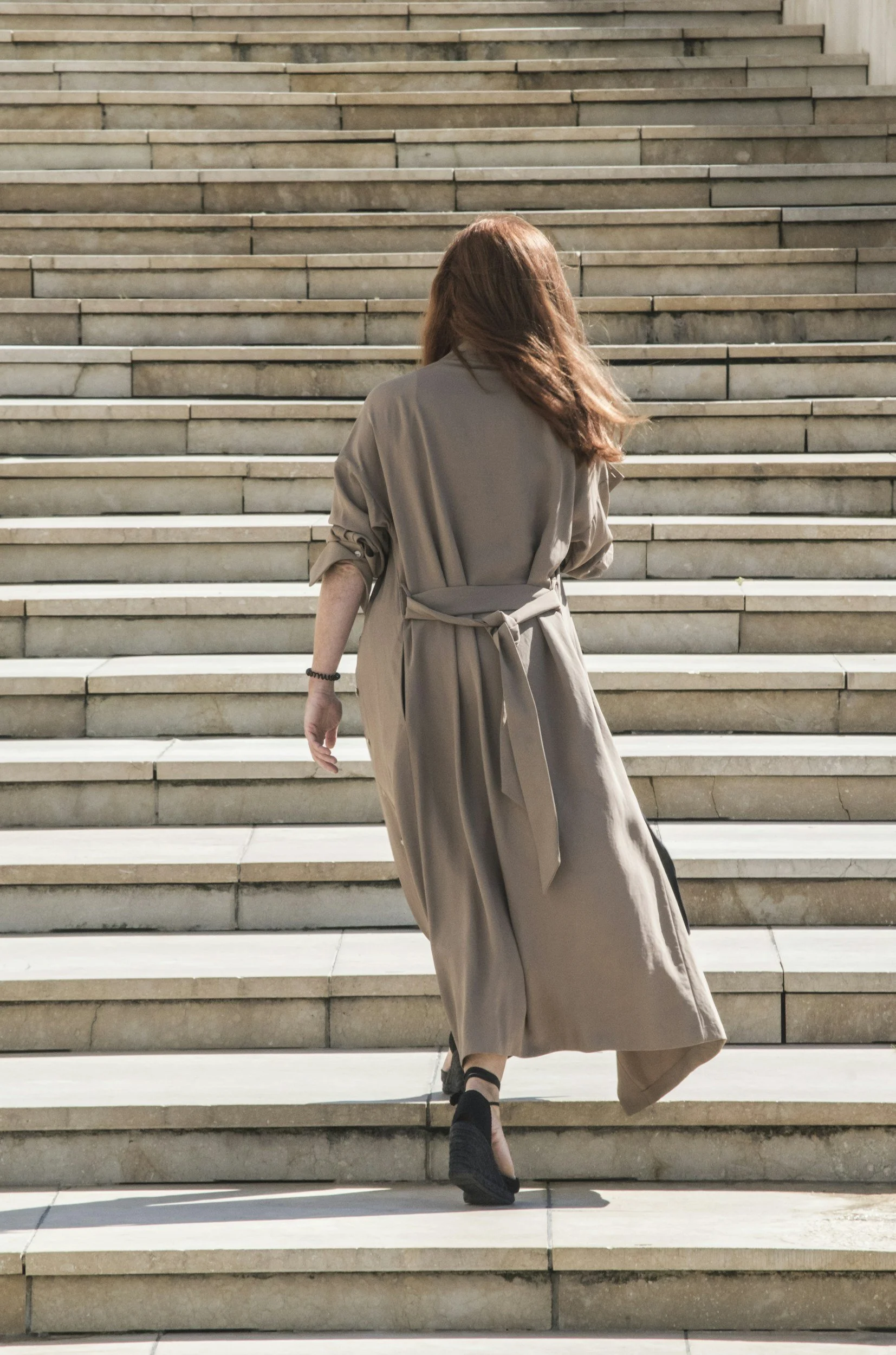 A woman with long red hair wearing a tan trench coat walking up stone stairs outdoors.