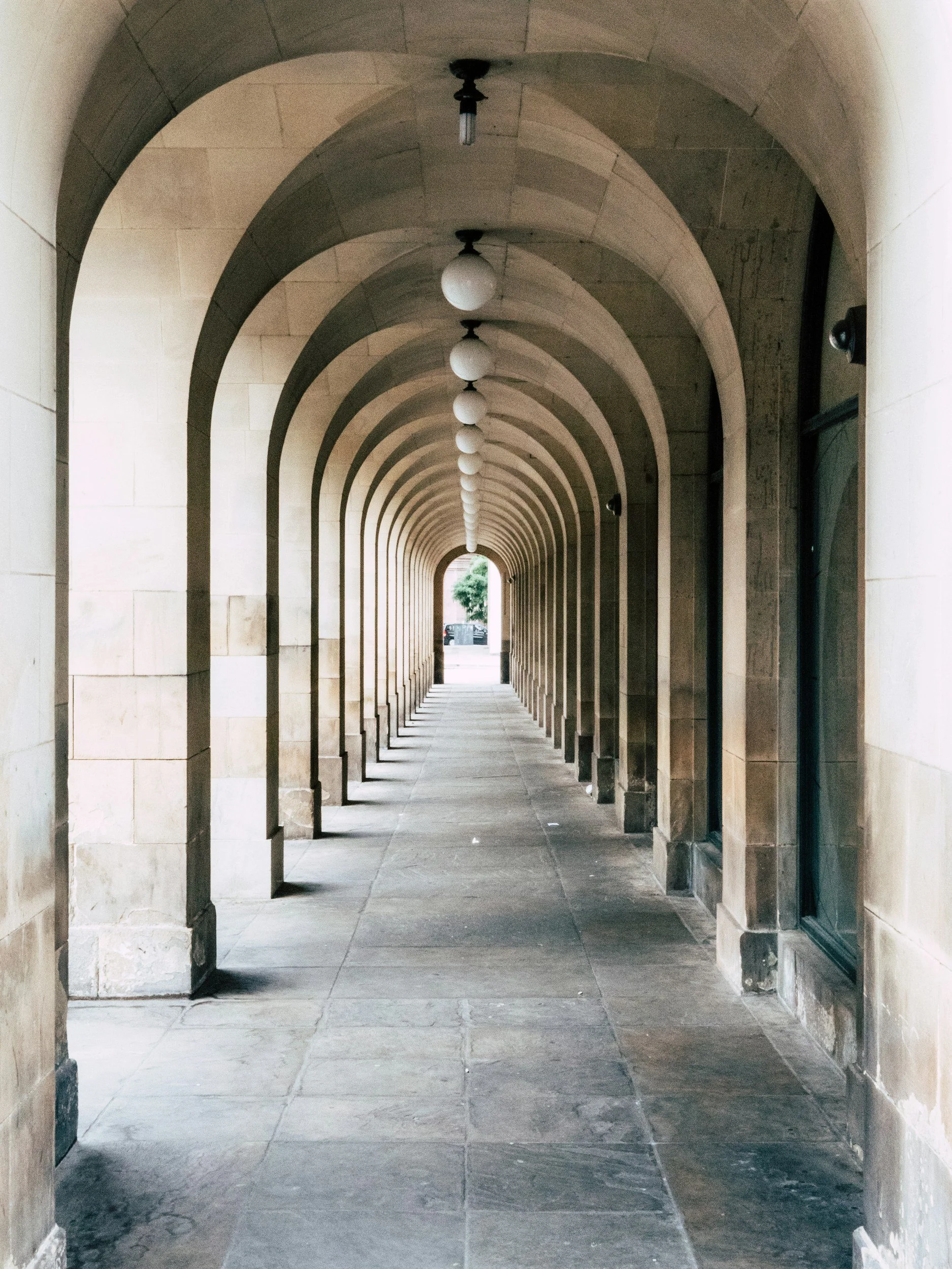 Long stone corridor with arched doorways, hanging lights, and a view of trees and cars at the end.