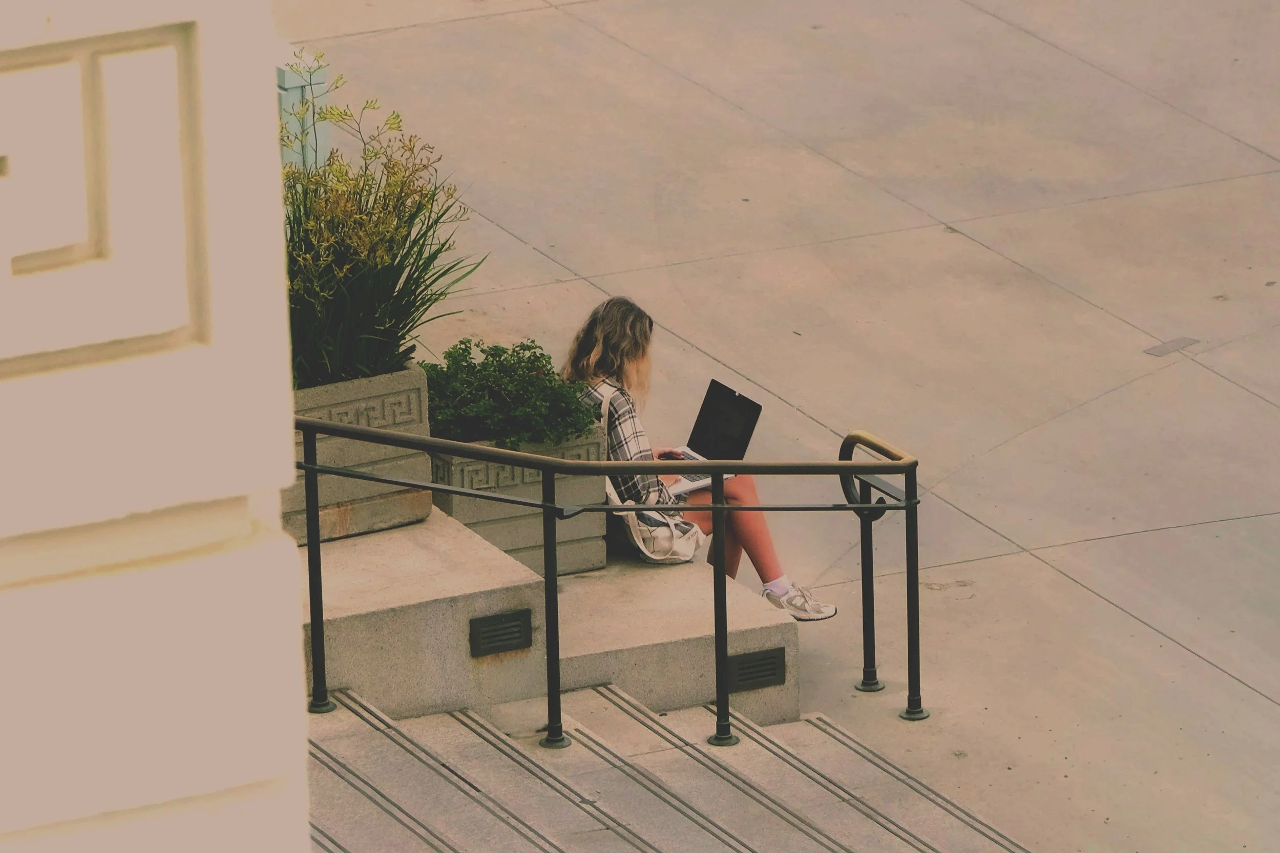 A young woman sitting on concrete steps outside a building, working on a laptop. She has shoulder-length hair, wears a plaid shirt, shorts, and sneakers, with a backpack beside her. There are plants and a railing nearby.