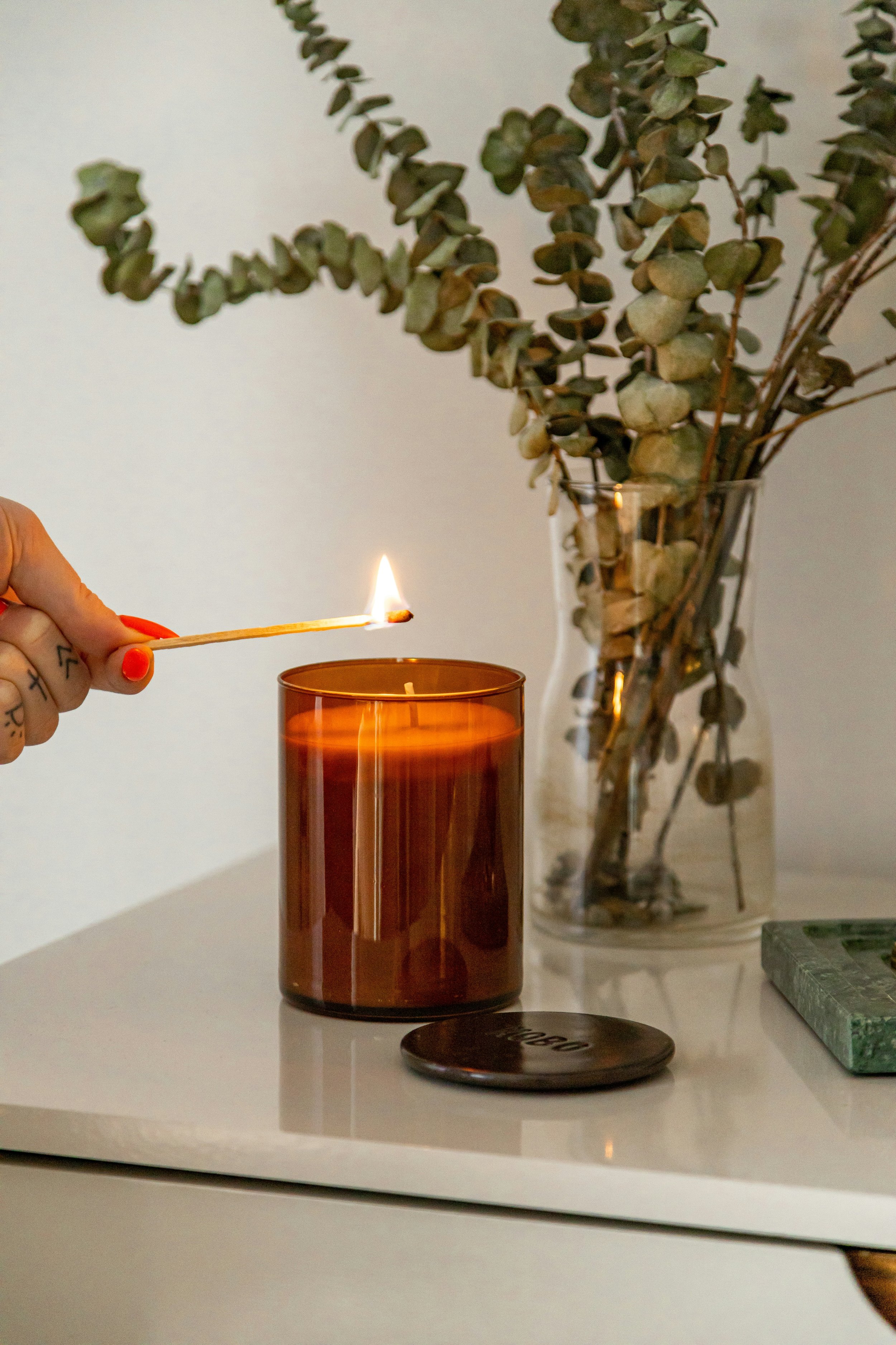 Person lighting a candle with a match on a white table, with a vase of dried eucalyptus branches in the background.