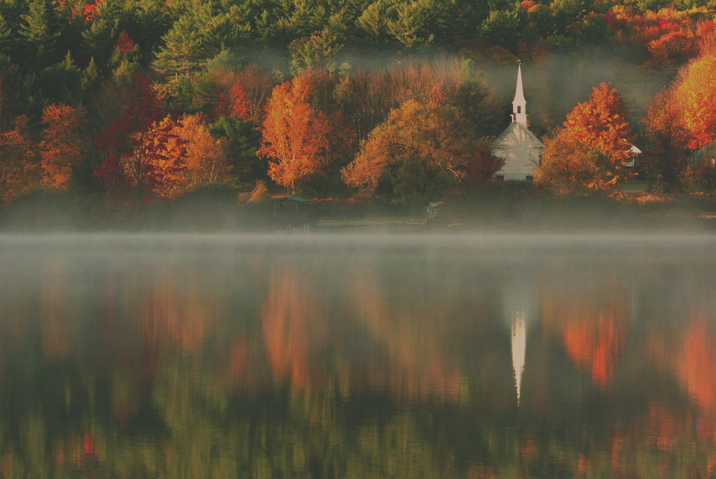 A lake with a mist over its surface, reflecting colorful autumn trees and a white church with a steeple on the opposite shore.