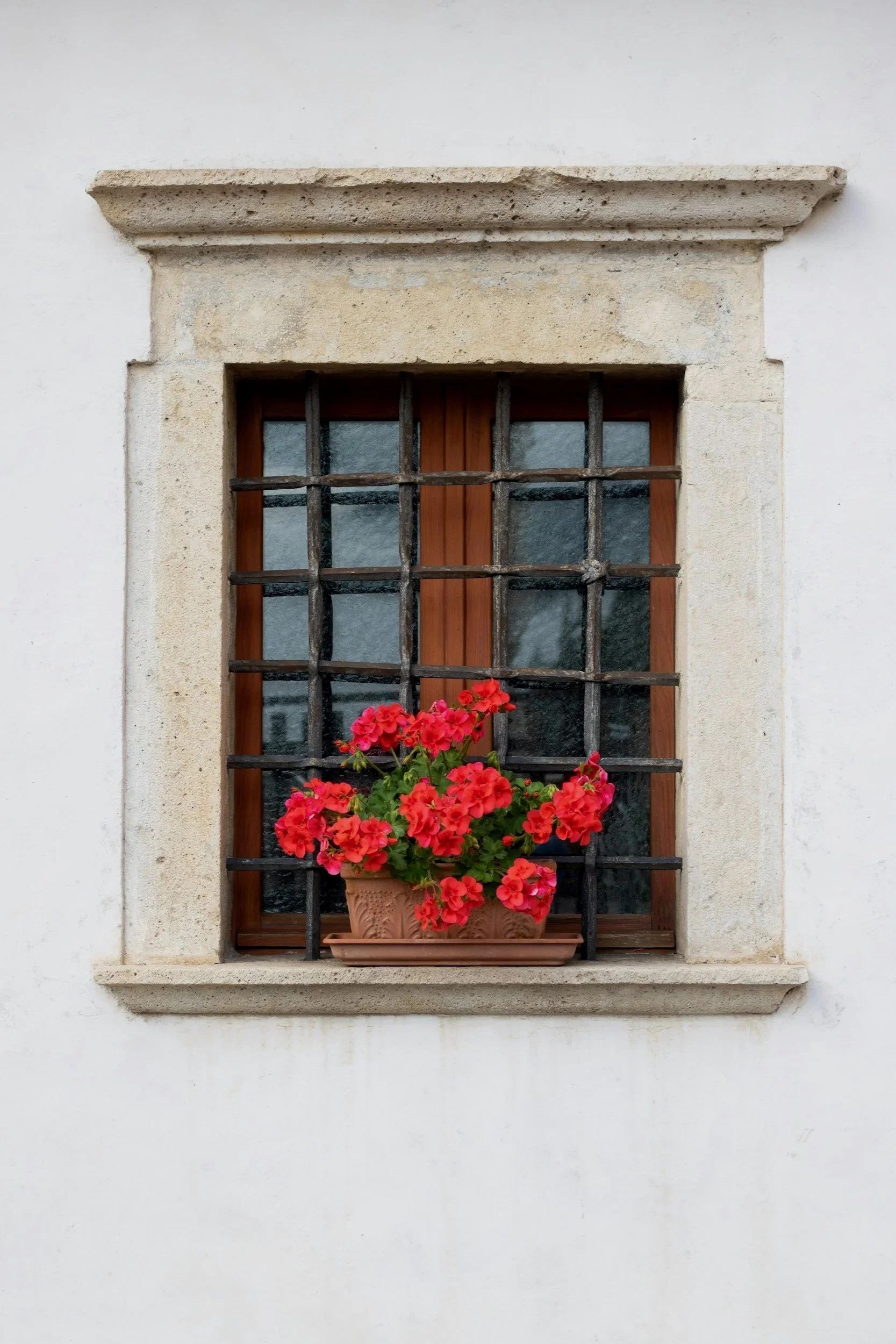 A window with wooden framing, black iron bars, a stone sill and frame, and a flowerpot of red flowers on the sill, set into a white wall.