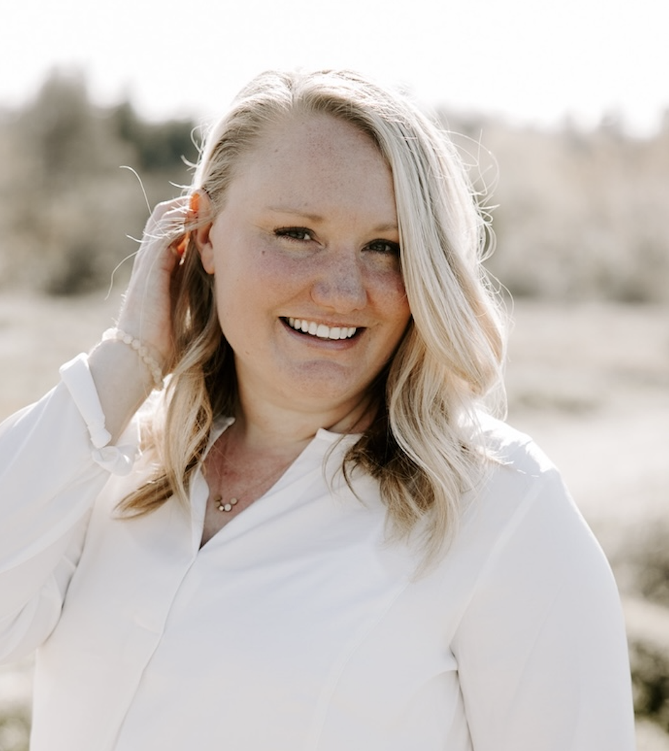 A smiling woman with blonde hair, wearing a white blouse, standing outdoors in bright sunlight with a blurred natural background.