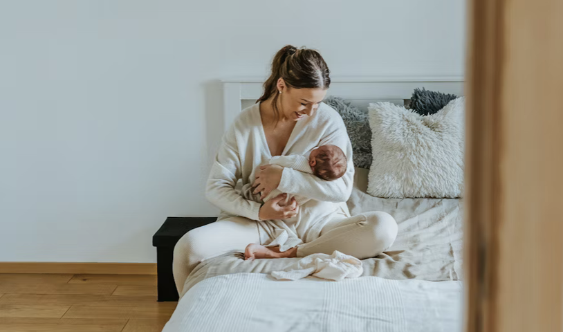 A woman sitting on a bed, breastfeeding a newborn baby, smiling and looking at the baby.