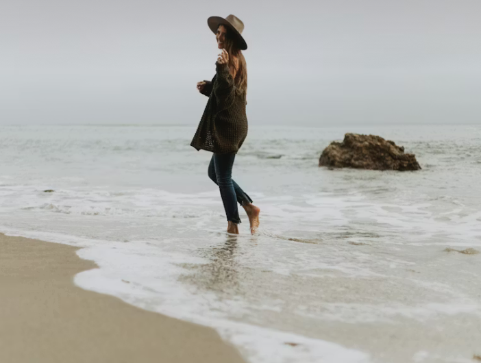 Woman in a hat and coat walking in shallow ocean water at the beach on a cloudy day.