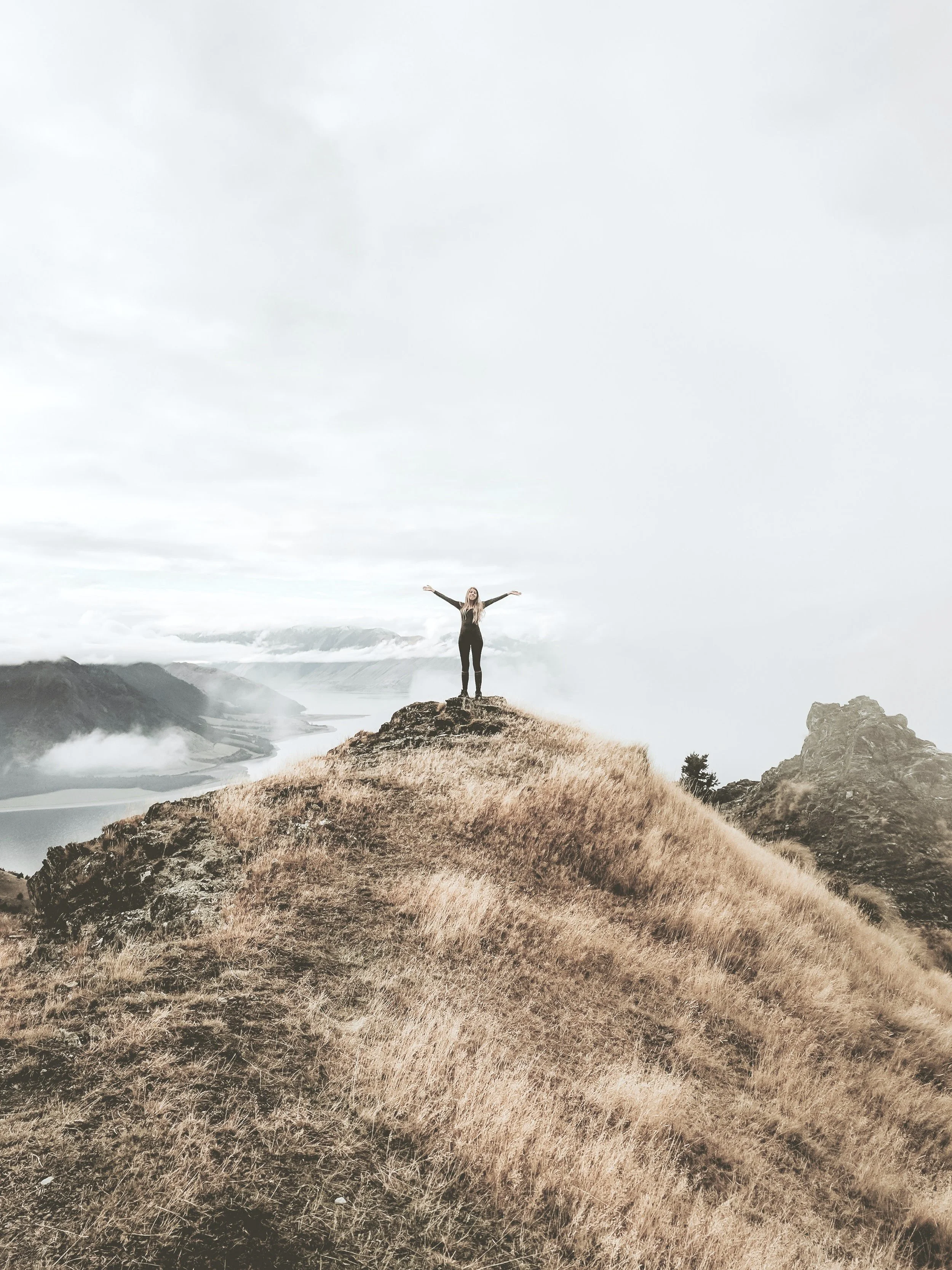 Person standing on a grassy hilltop with arms raised, surrounded by clouds and mountain scenery.