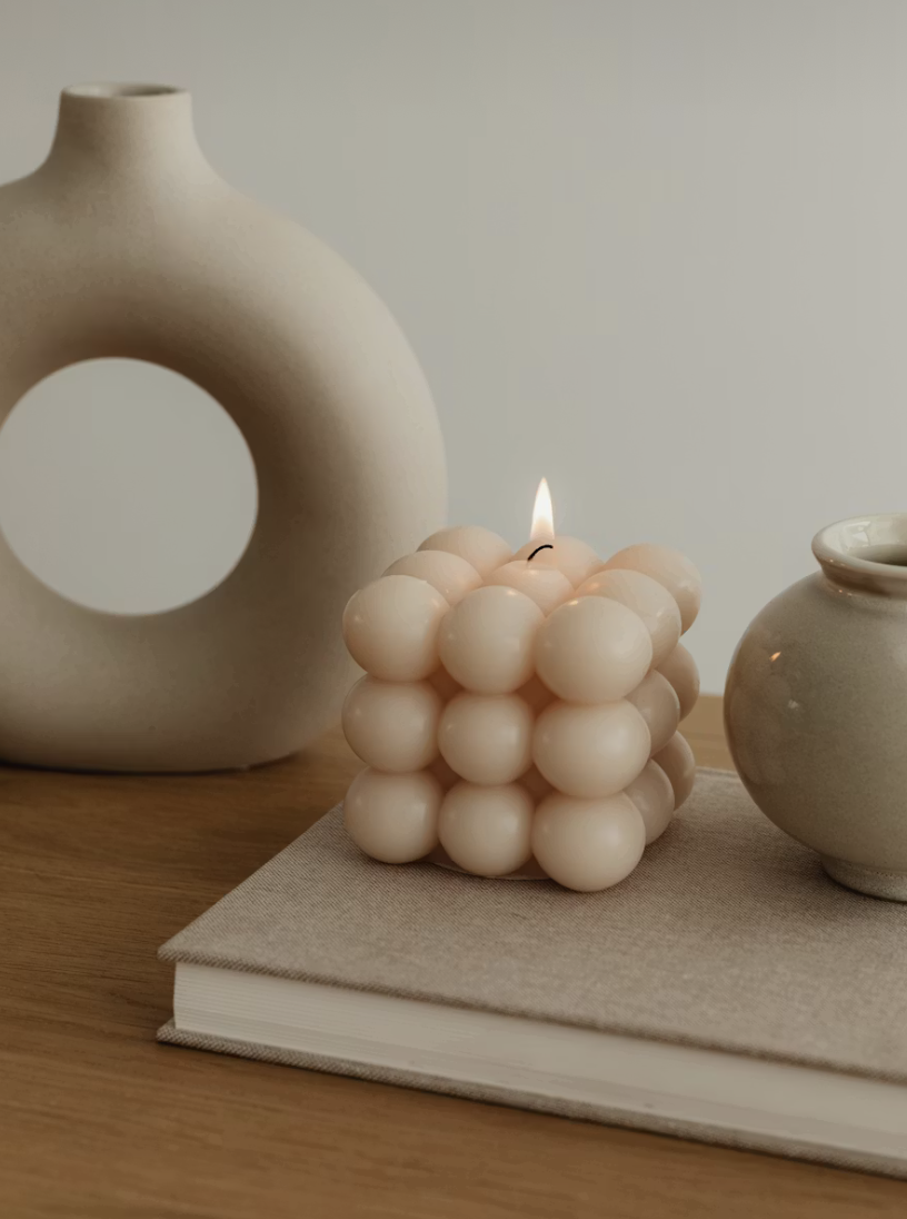 Decorative scene with beige spherical candle lit on a closed book, flanked by a large ceramic vase with a circular cutout on the left and a small ceramic jar on the right, on a wooden surface against a plain wall.