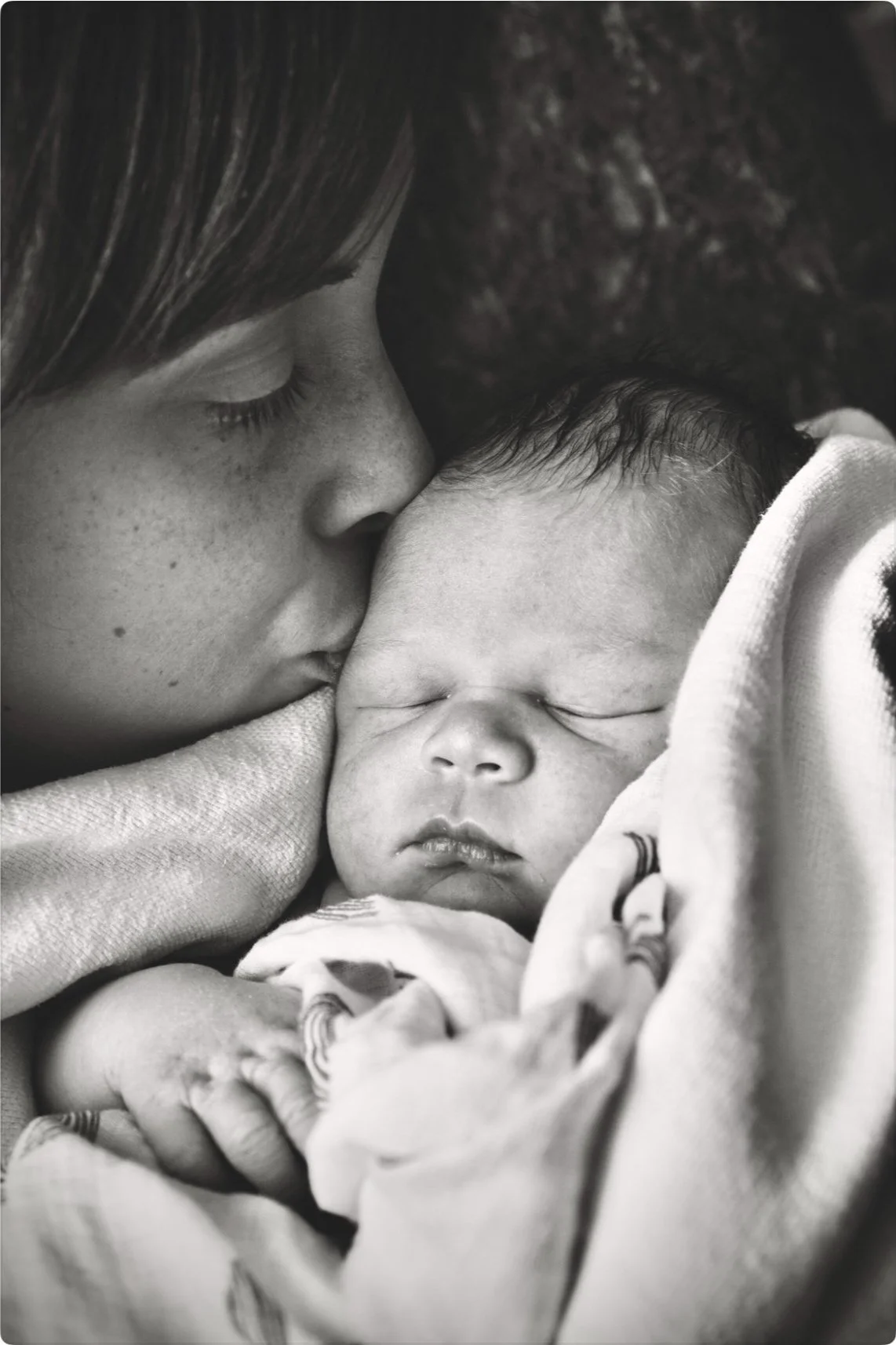 Black and white photo of a person kissing a sleeping baby wrapped in a blanket.