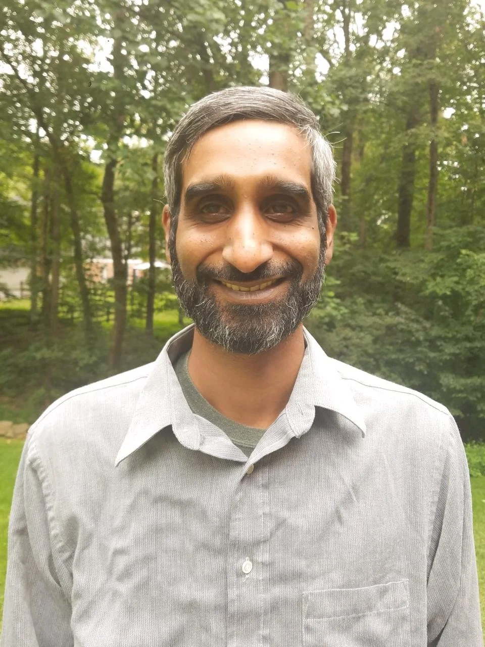photo of man outside, green trees behind him