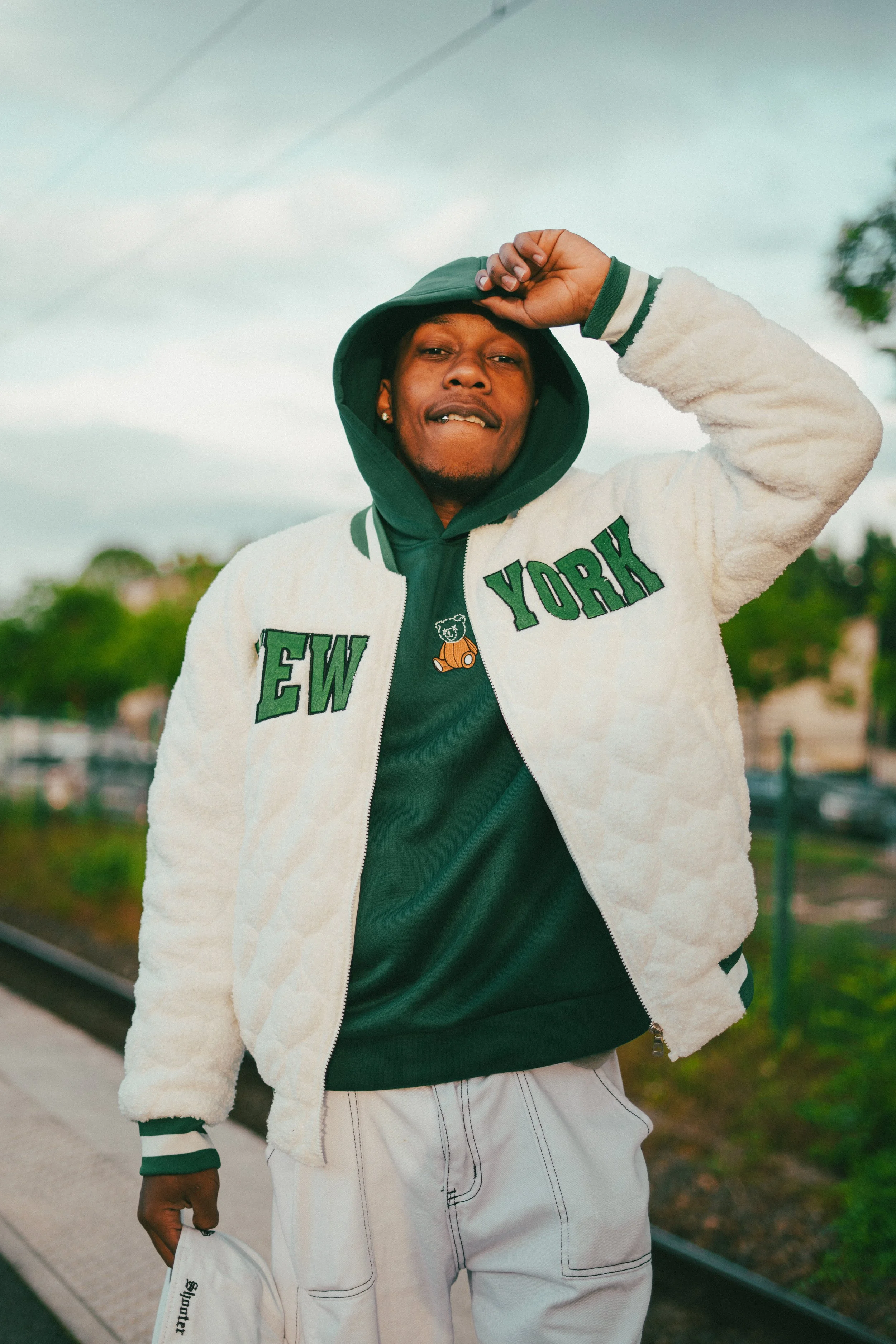 Jeune homme portant une veste blanche, un hoodie vert, et tenant une casquette blanche, dehors lors d'une journée nuageuse.