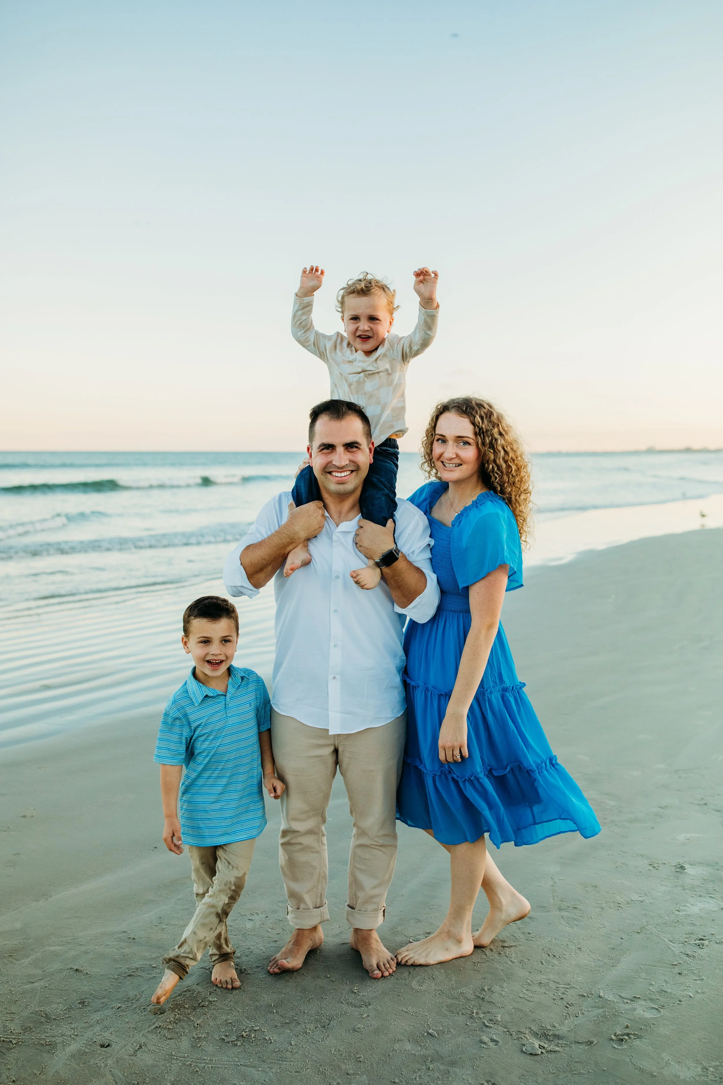 A happy family of four standing on a beach, with a father carrying his young son on his shoulders, a mother in a blue dress, and two boys, one in a striped shirt, all smiling and enjoying the ocean scenery at sunset.