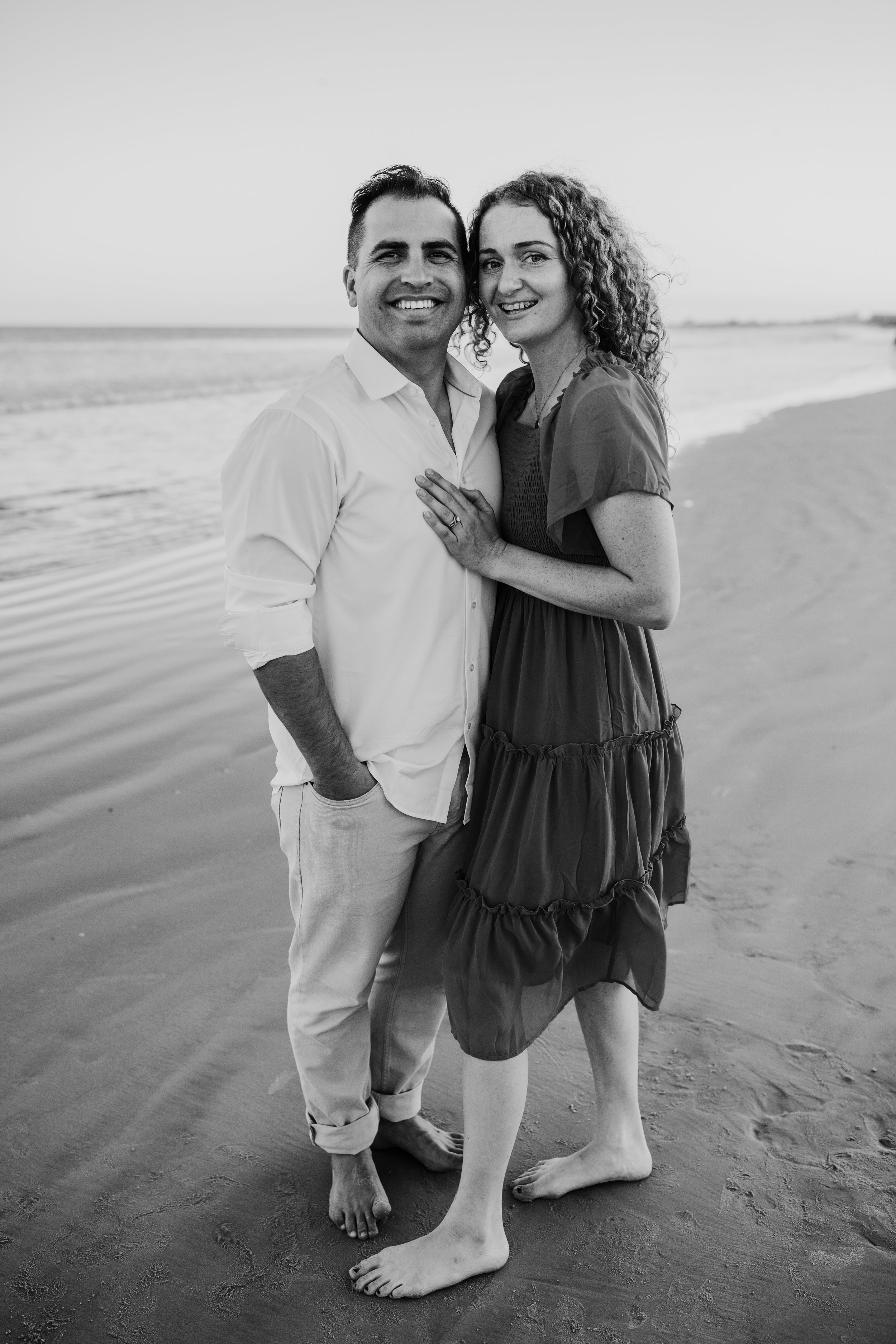 A black and white photo of a smiling couple standing on a beach. The man has short dark hair, is wearing a light button-up shirt with rolled-up sleeves and light pants with bare feet. The woman has curly hair, is wearing a dark dress with short sleeves, and is barefoot. They are standing close together, with the woman's hand resting on the man's chest, near his heart.