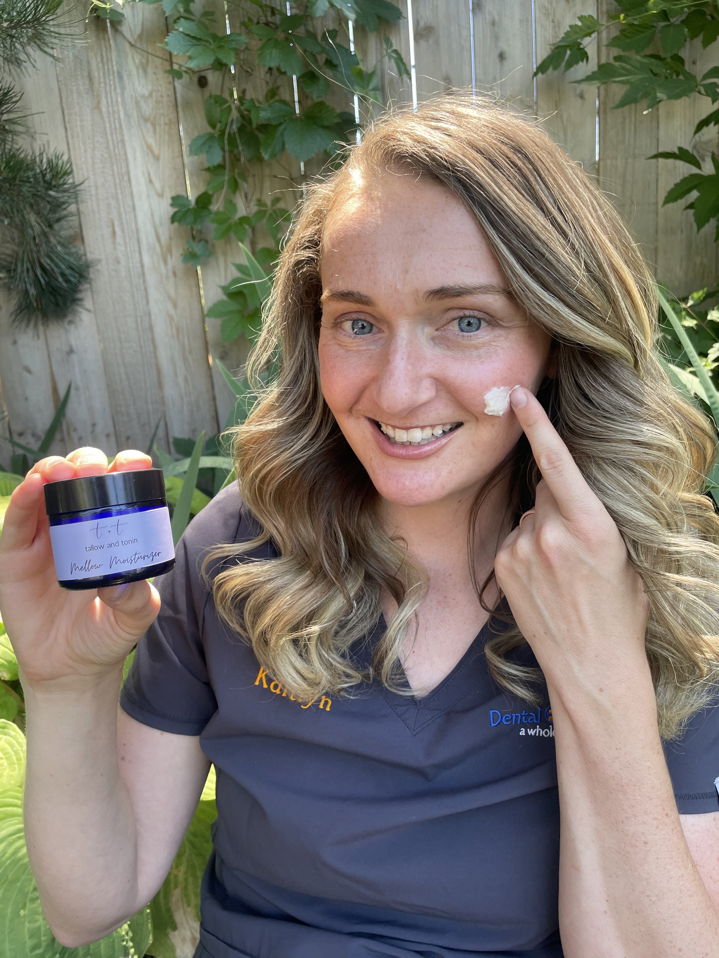 A woman with blonde wavy hair and blue eyes smiling outdoors. She is pointing to a small amount of cream on her cheek and holding a blue jar labeled Mellow Moisturizer In the background, there is a wooden fence with green leafy plants.
