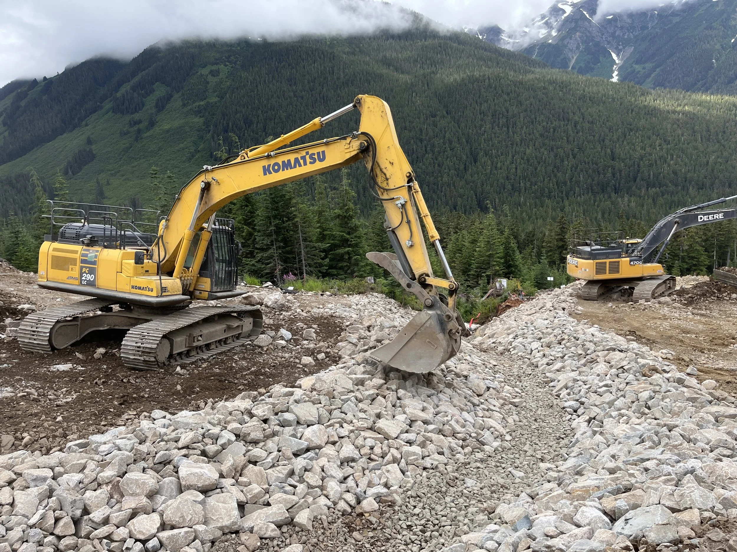 Construction site with two yellow excavators working in a mountainous area with forested slopes and cloudy sky.