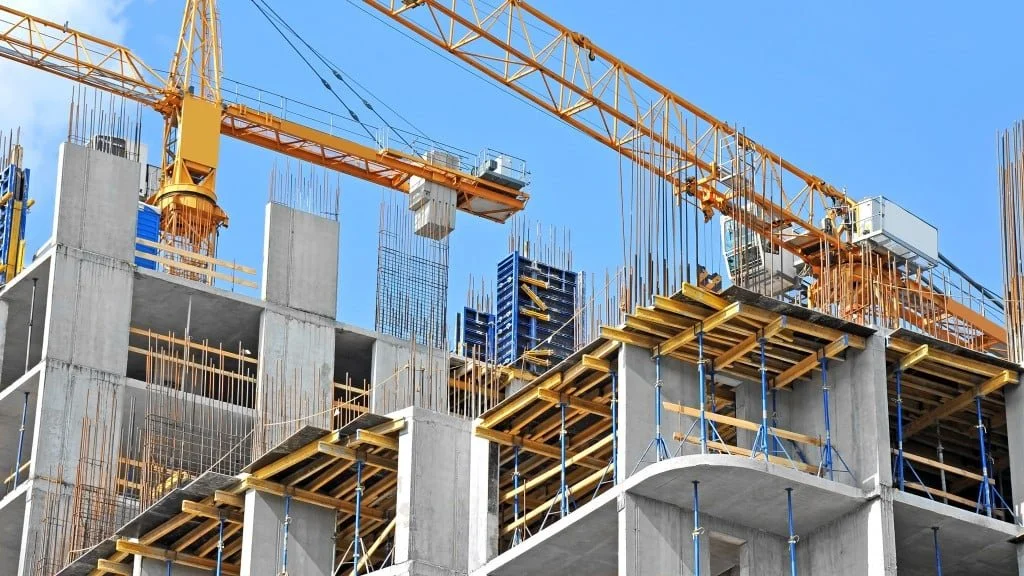 Construction site with concrete building framework, wooden formwork, steel reinforcement bars, and a yellow tower crane against a blue sky.