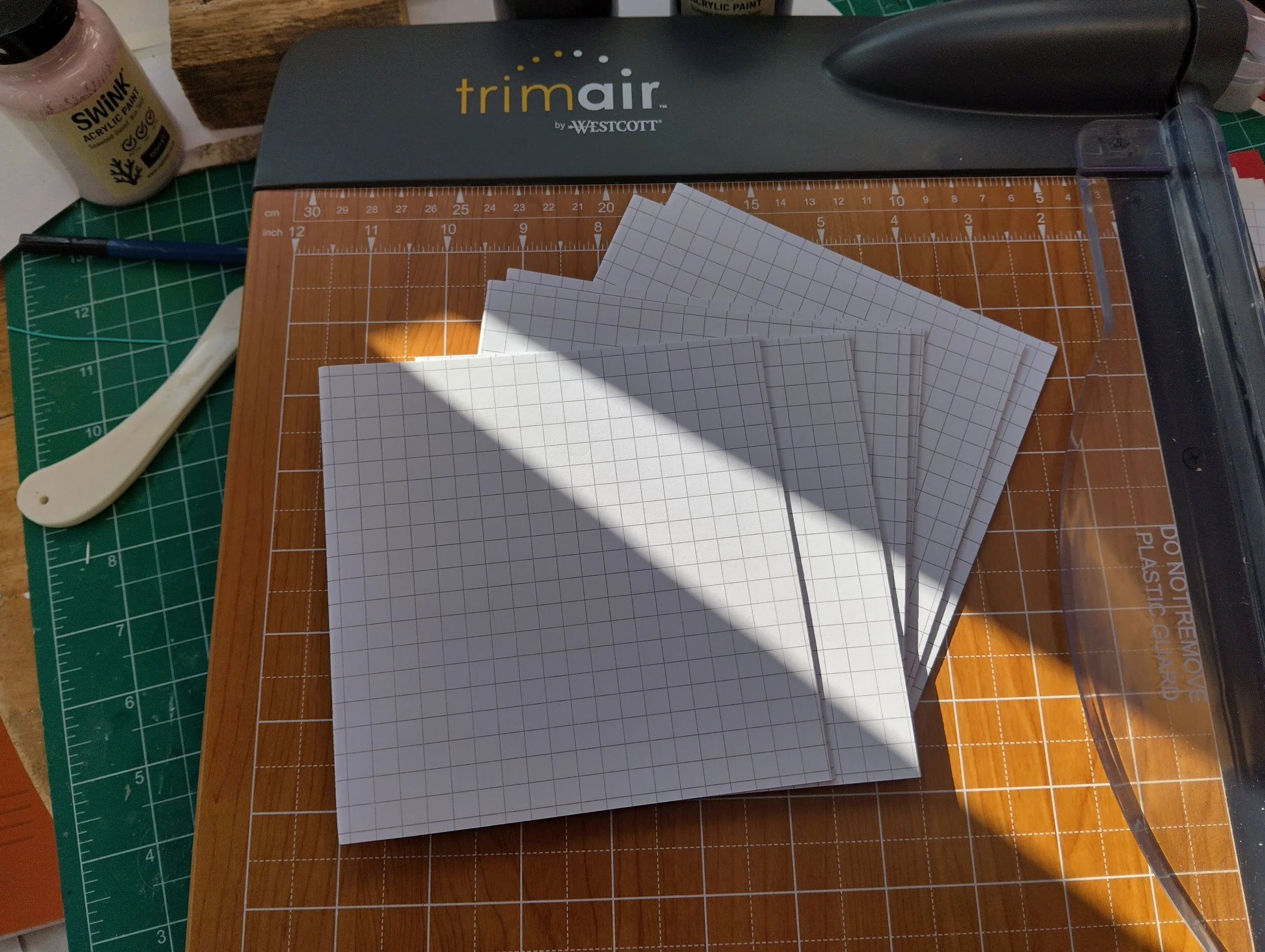 A workspace with paper, a paper cutter, a marker, a paint bottle, and cutting mats on a wooden table.