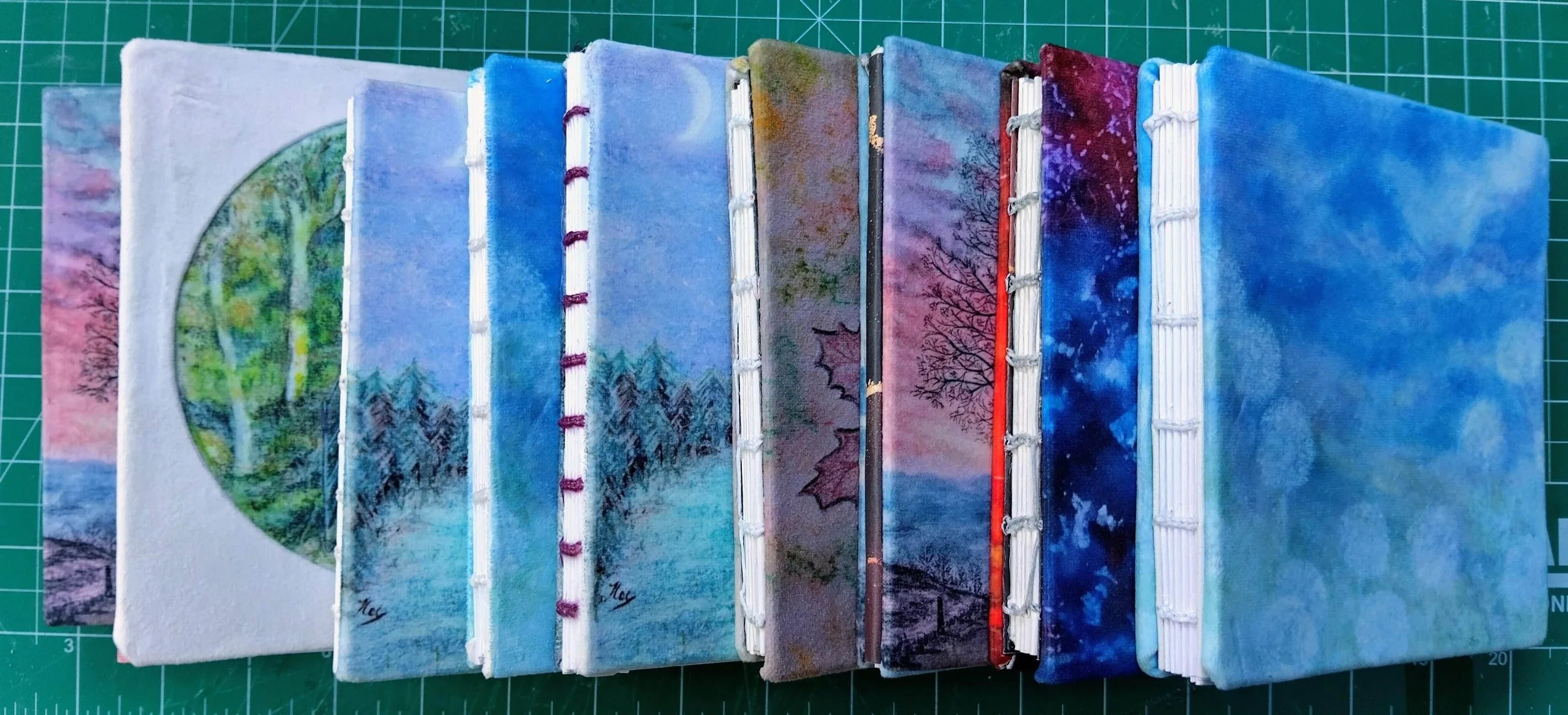 A collection of handbound notebooks with nature and landscape paintings, arranged on a green grid cutting mat.