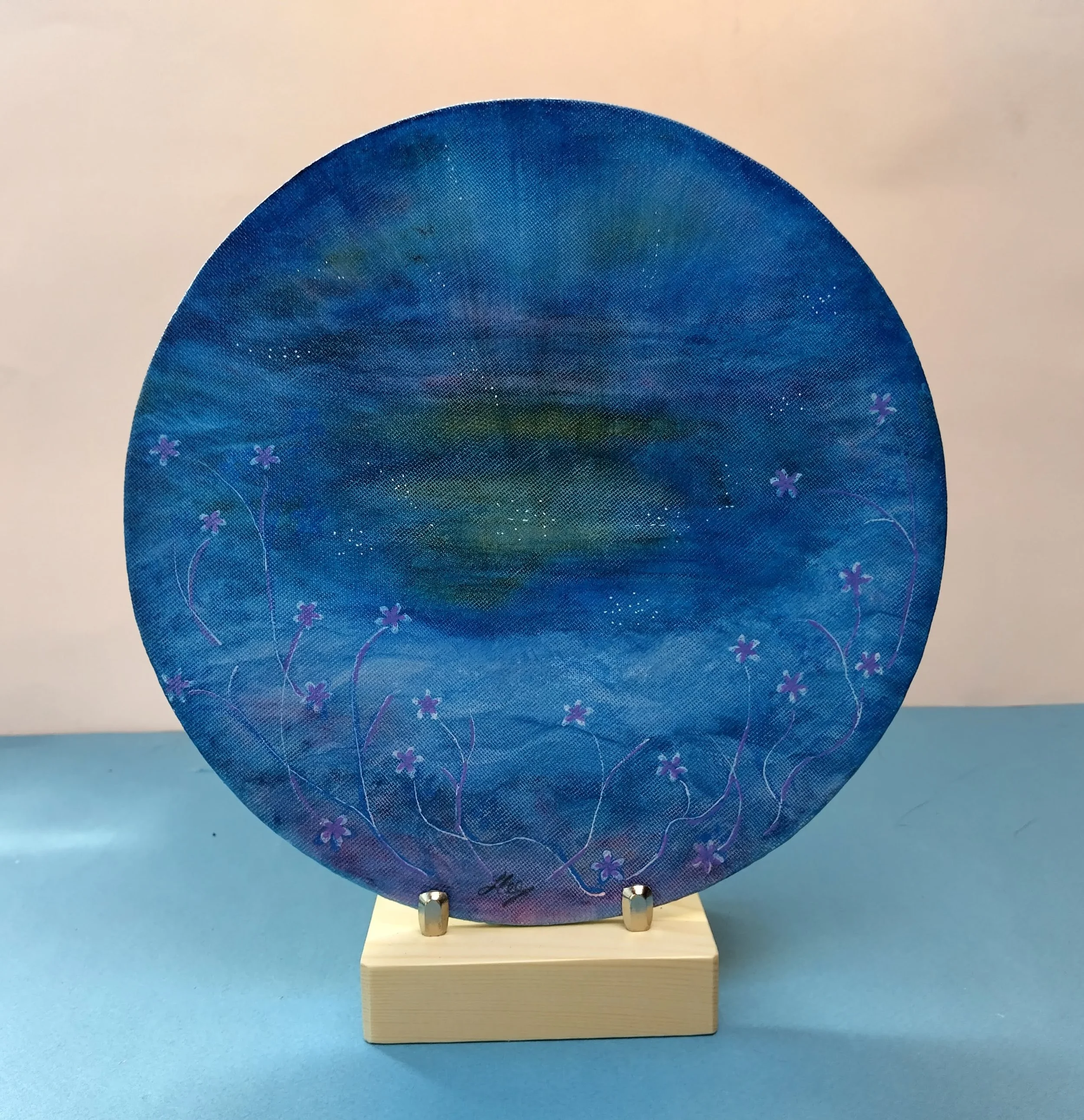 A semi abstract circular painting in blues, with a warm yellow at its heart and cool coloured flowers in the foreground