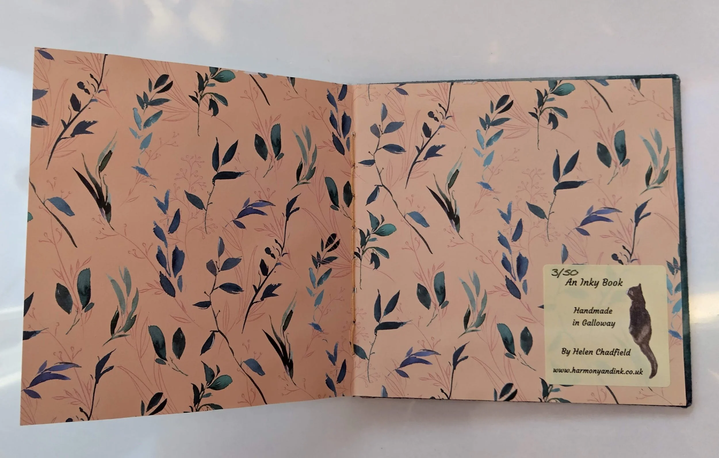 Open pink and beige watercolor-style notebook with black and green leafy plant illustrations, labeled as '3/50 An Inky Book Handmade in Galloway' by Helen Chadfield with a website URL.