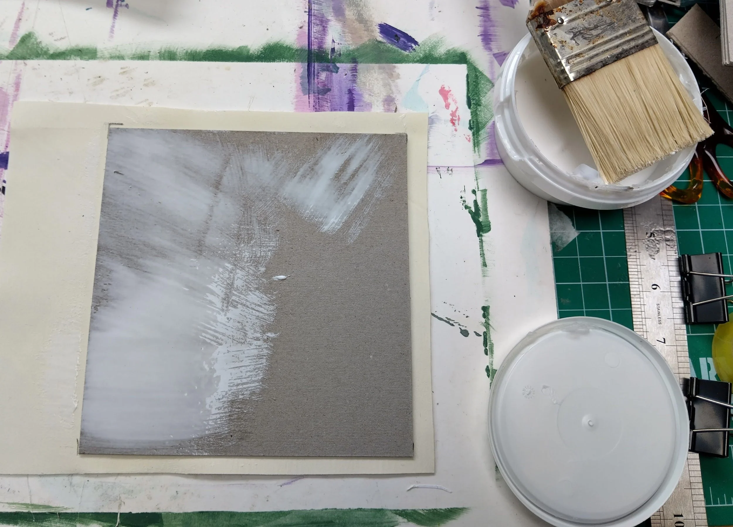 Art workspace with paper and glue, a paintbrush in a White container, and a ruler on a digital cutting mat.