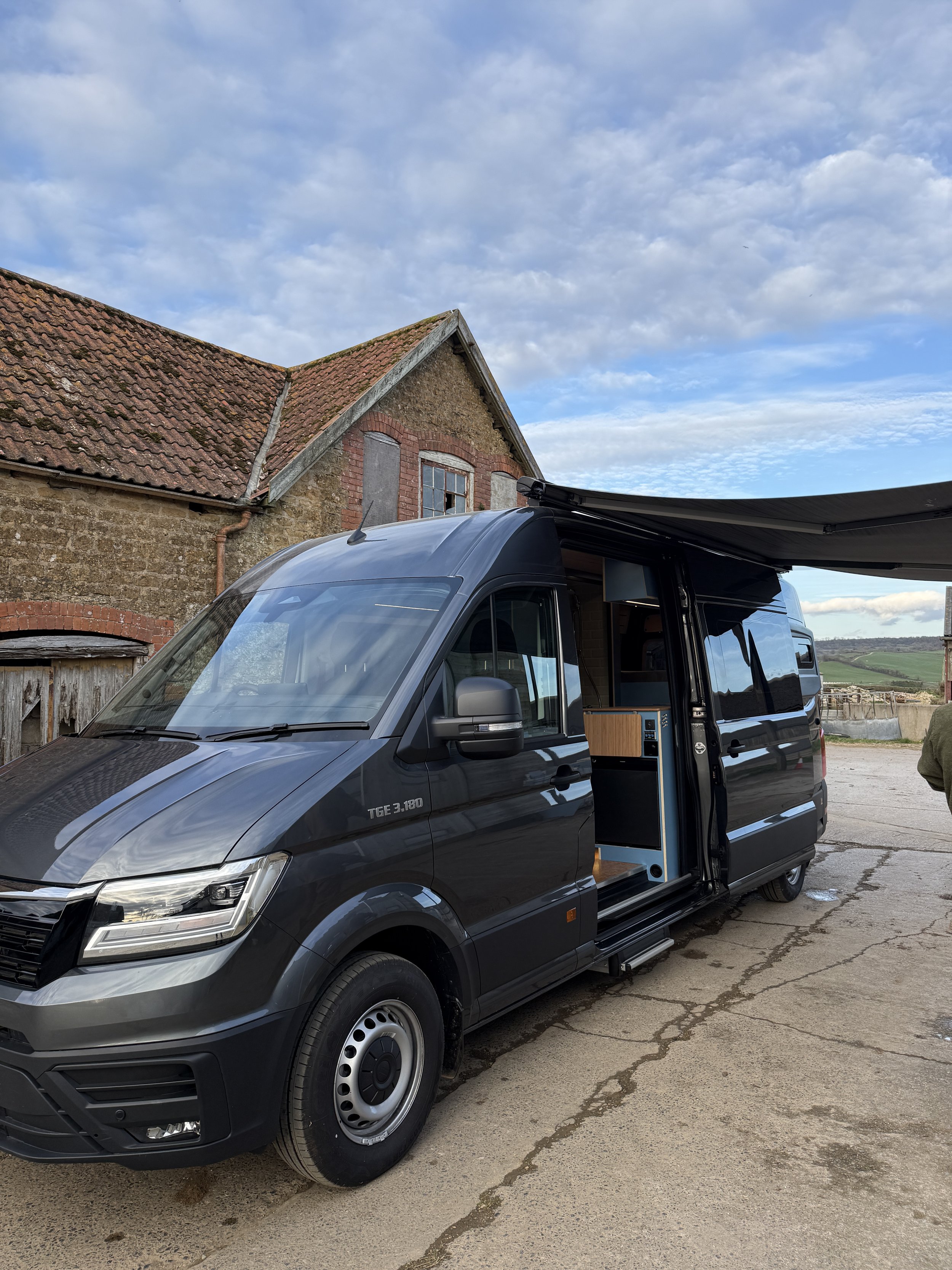 A black camper van with an open side door and an extendable awning parked on a cracked concrete surface in front of an old rustic building with a tiled roof and brick accents. The sky is partly cloudy with patches of blue.