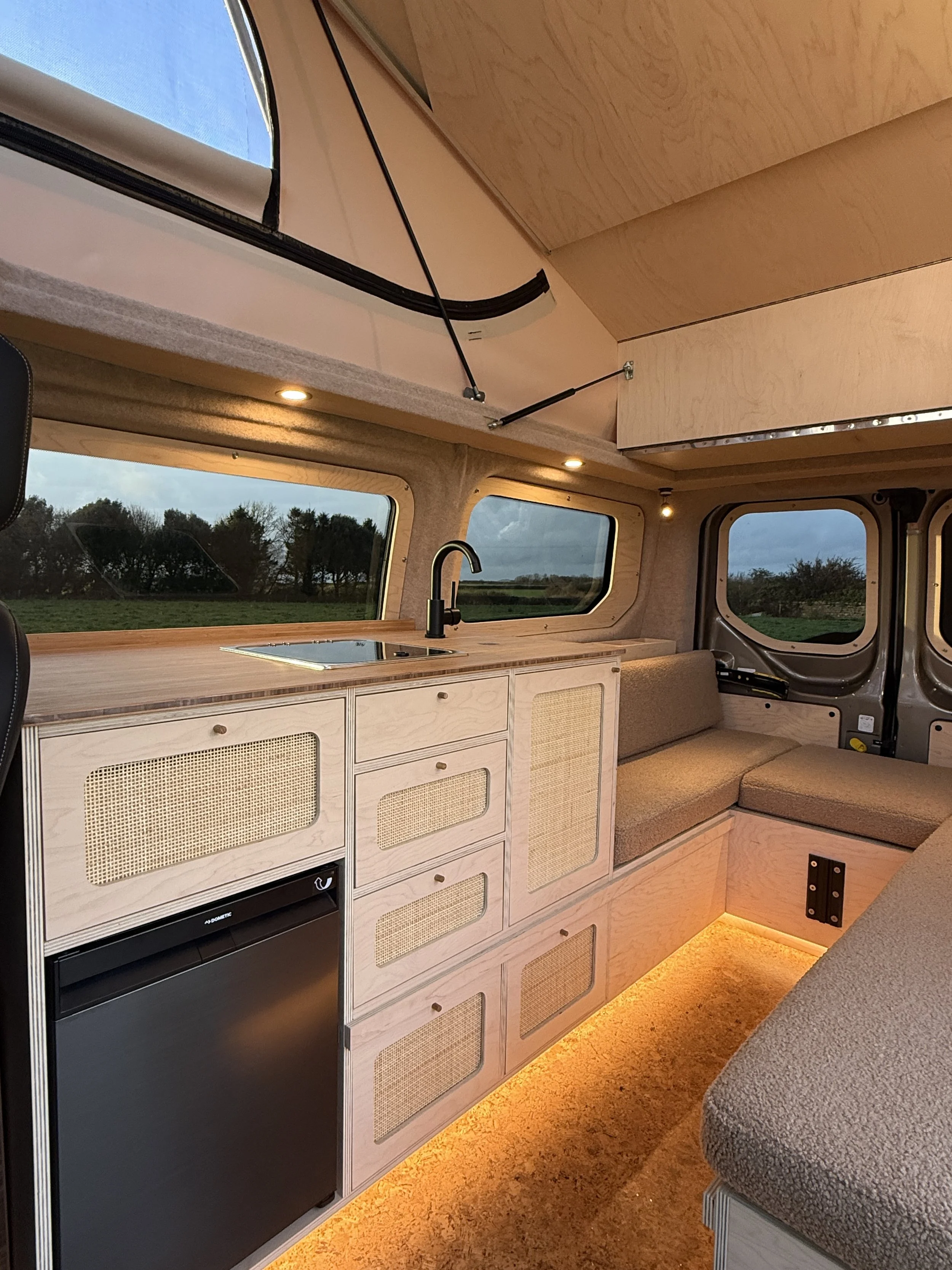 Interior of a camper van with a kitchenette, beige cabinets, a small sink, a window view of a grassy field, and a beige cushioned seating area, illuminated with warm LED lighting.