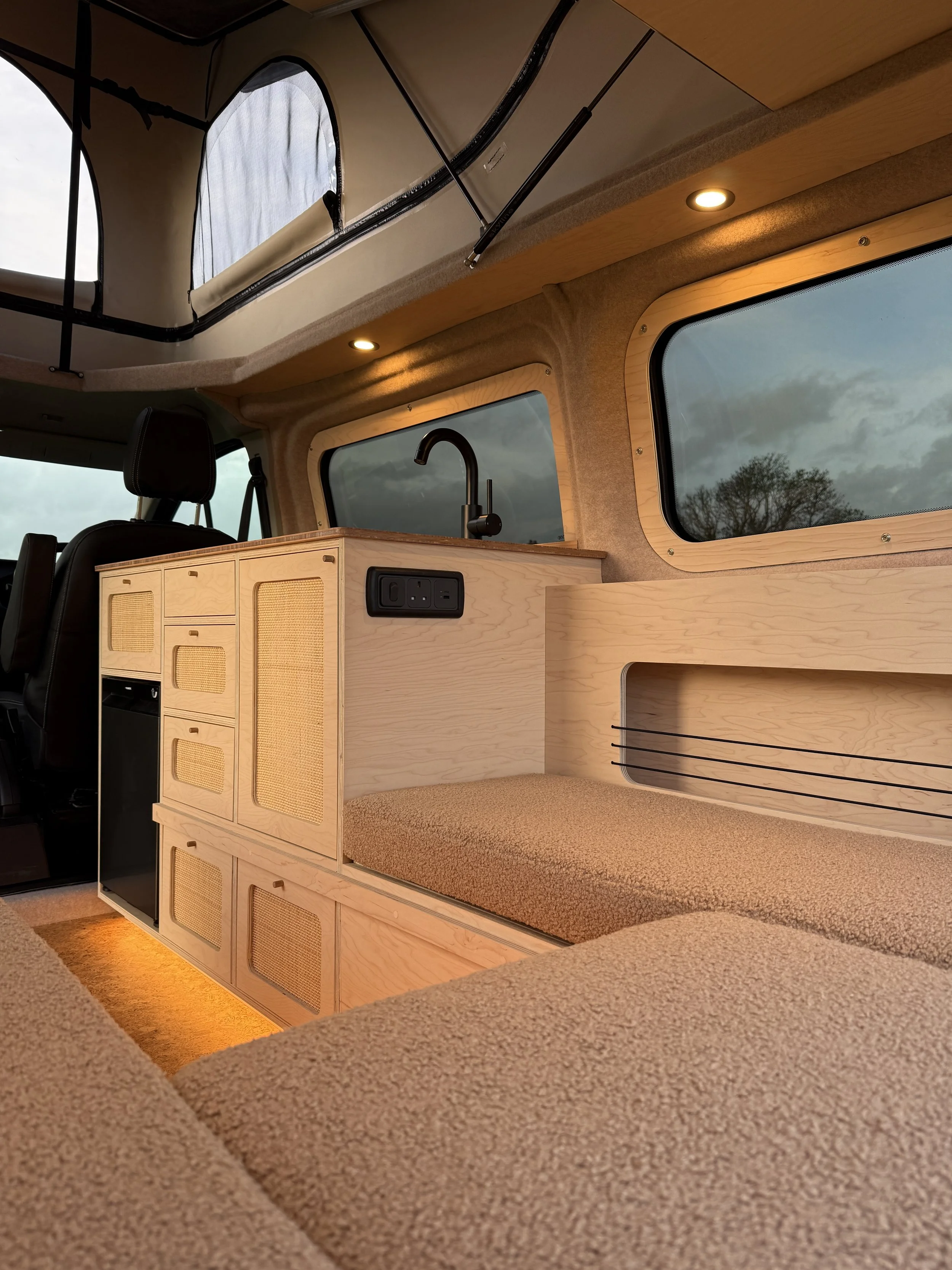 Interior of a camper van showing a wooden kitchen area with a small refrigerator, a countertop, a built-in bench with storage underneath, a window, and a ceiling with skylights and recessed lights.