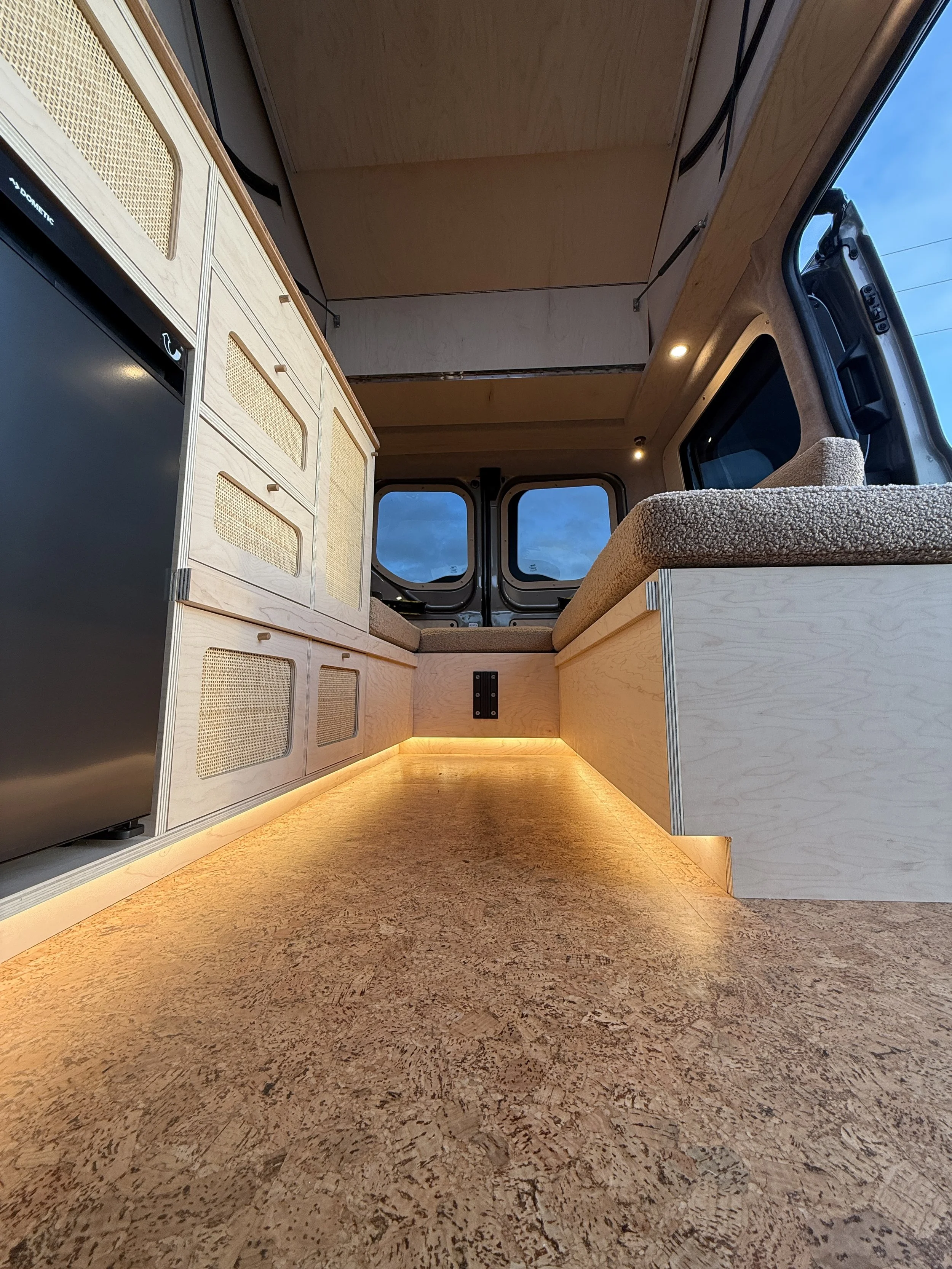 Interior of a camper van with wooden cabinets, fabric seating, and warm under-cabinet lighting.