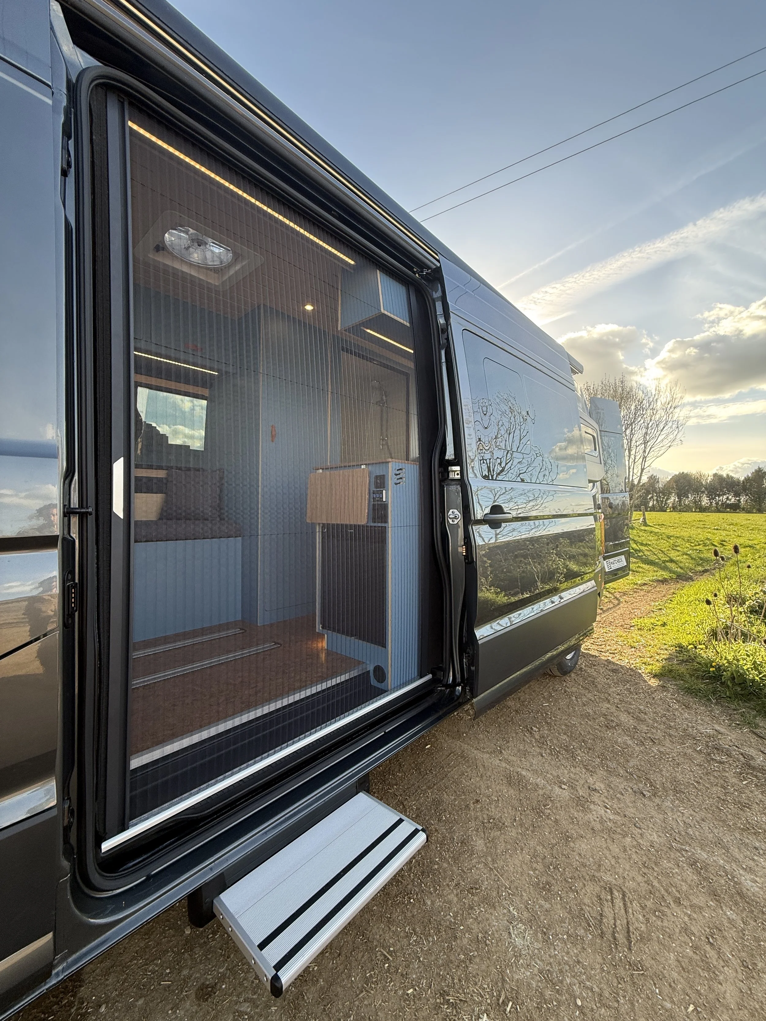 The image shows the side of a black camper van with an open sliding door, revealing the interior which has wood flooring, blue walls, and furniture. The camper is parked outdoors on a dirt path, with a grassy field and trees in the background, under 