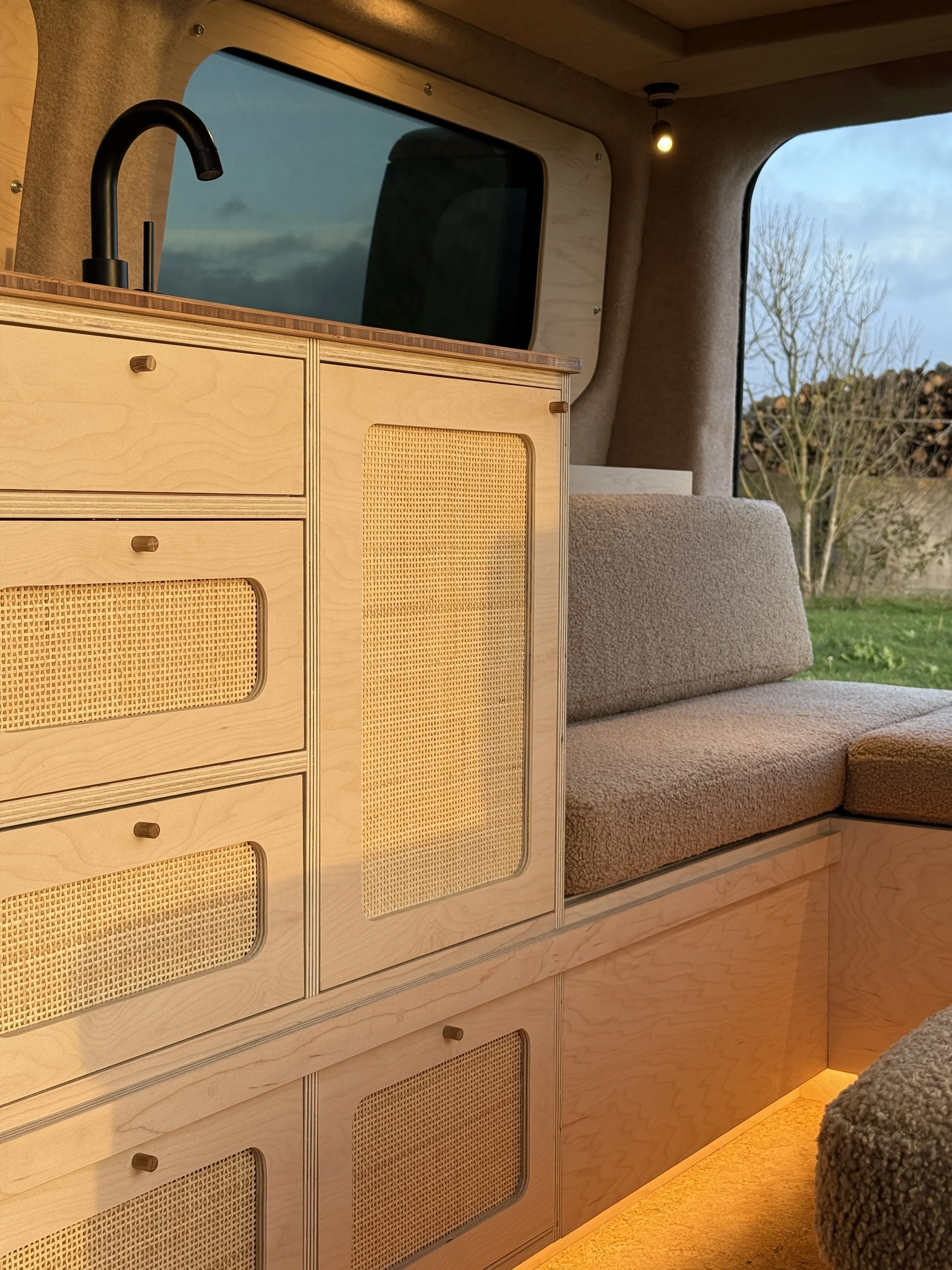 Interior of a camper van with light wooden cabinetry, a black sink faucet, beige cushioned seating, and a window showing a view of trees and a cloudy sky outside.