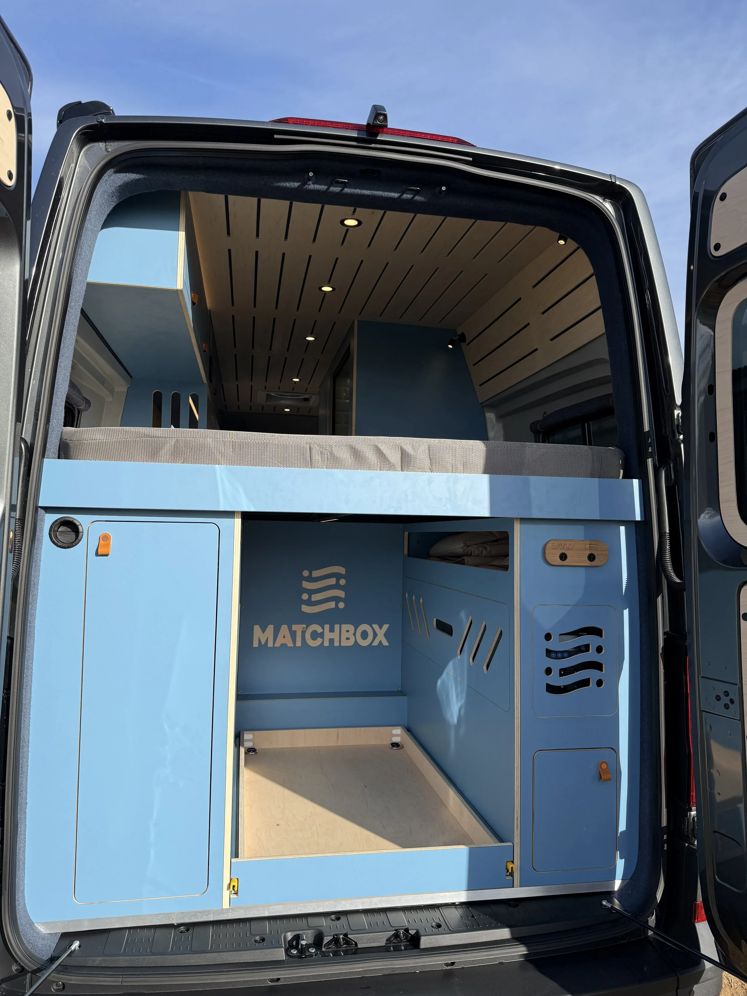 Inside of a camper van with a blue custom storage unit labeled 'MATCHBOX' and a living area with a bed and wood-paneled ceiling.